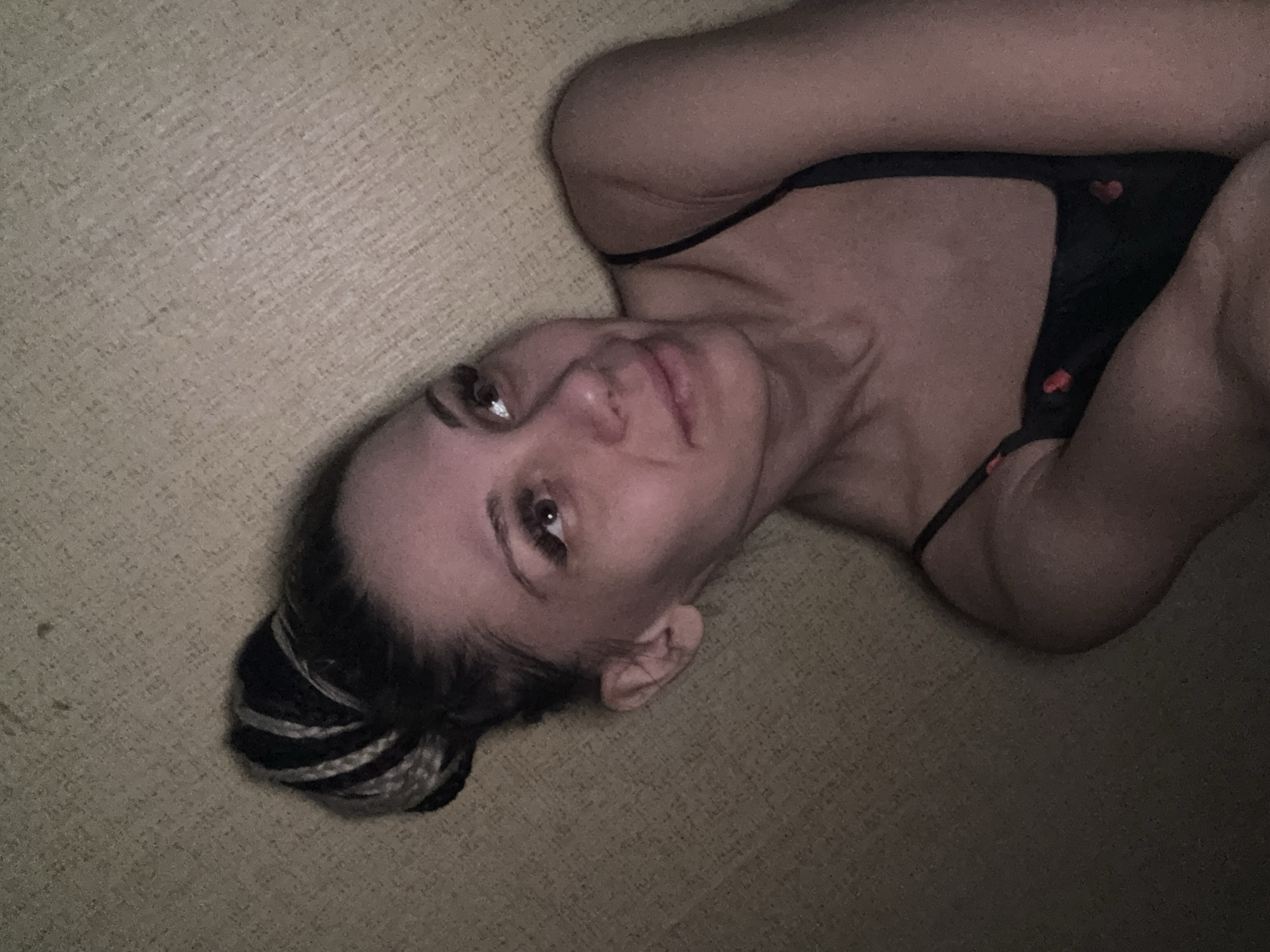 A person lies on a carpeted floor. The person is wearing a black camisole with red hearts. Their hair is in a bun with black and white braids. The location is not discernible from the image.