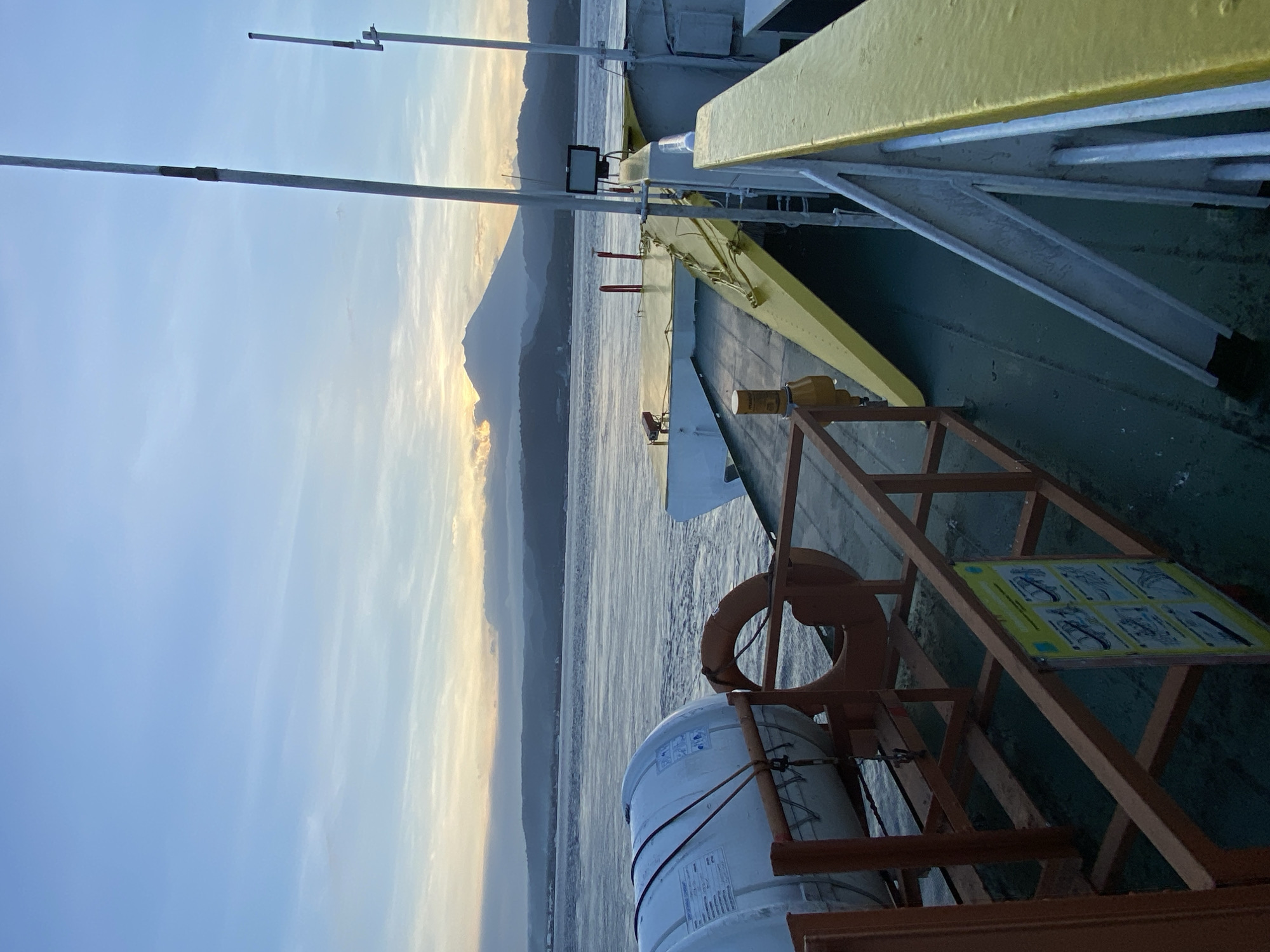 The image shows a view from the deck of a vessel. Visible on the deck is safety equipment including a life ring and a white cylindrical object secured by ropes within a metal frame. A yellow-painted railing is also present. In the background, land can be seen across a body of water with a mountain range visible along the horizon illuminated by the sunset. The sky is light blue with streaks of white and yellow near the horizon. The image is taken during daylight hours. Location is Unknown, Indonesia.