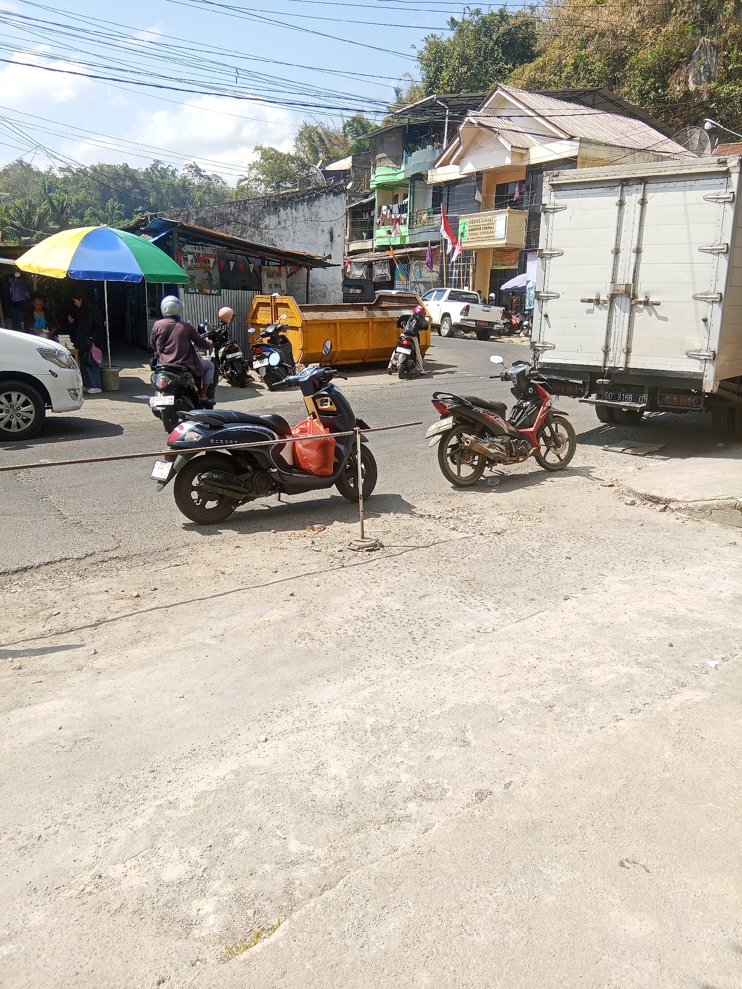 The image depicts a street scene in Makale, Indonesia, likely during the daytime under a clear sky. The foreground is dominated by a dusty, unpaved area with some scattered debris. A portion of an asphalt road is visible, and on it are parked several motorcycles. One dark-colored scooter, with an orange bag on its rear, is prominently featured in the middle ground. Another red and black motorcycle is parked to its right. Further back on the road, two more motorcycles are visible, one with a rider wearing a helmet.
To the left of the road, a white car is partially visible. Adjacent to it, a market stall is set up with a colorful umbrella providing shade. Several people are seen near the stall, with some appearing to be customers and vendors. Behind the stall, a yellow dumpster is situated.
Across the road, a row of buildings can be seen. Some are low-lying structures with awnings, while others are multi-story buildings. One building has a sign with Indonesian text and flags displayed. On the right side of the image, the back of a large white truck with a container is parked, obscuring some of the buildings behind it. The truck's license plate is visible. In the background, a hillside covered in vegetation rises behind the buildings. Power lines are strung across the sky.