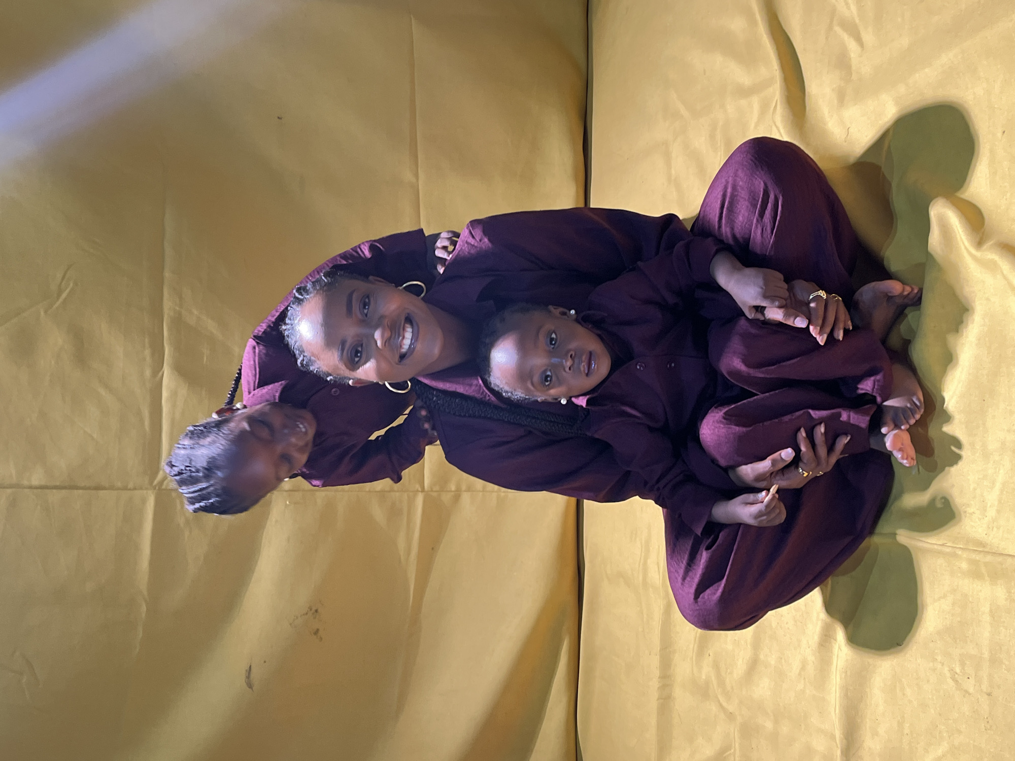In an indoor studio setting with a plain, crinkled, yellow backdrop, a Black woman and two Black children, likely her daughters, are posed. The woman is seated cross-legged, facing the camera with a warm smile. She is wearing a dark maroon collared shirt and matching wide-leg pants. Large gold hoop earrings adorn her ears. A younger child, also in dark maroon attire, sits on the woman's lap, looking directly at the camera with wide eyes. An older child, dressed in the same maroon outfit, leans in from behind the woman's left shoulder, looking down and to the left, with a playful expression. The older child has braided hair. The younger child has hair styled in small braids with beads. The woman's hands are resting on her lap, one holding the hand of the child on her lap. The scene is lit from above, creating shadows on the yellow backdrop. The overall impression is a family portrait with a focus on connection and warmth, possibly taken during a professional photoshoot in Jos, Nigeria.