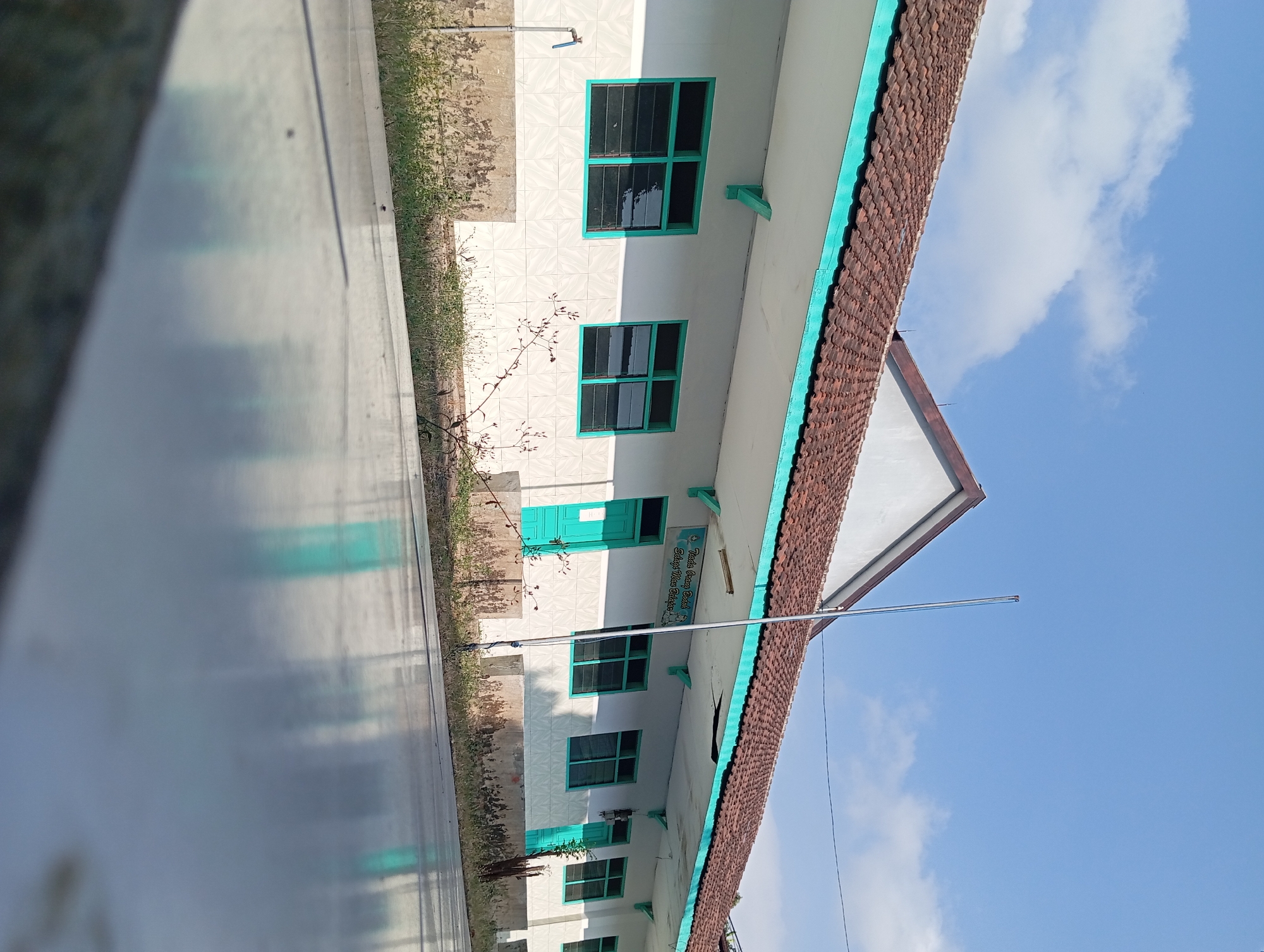 A low-angle, eye-level, full shot captures a white, single-story school building with a red tile roof under a bright blue sky with scattered white clouds. The building has several green-framed windows and green doors. A sign with Indonesian text is visible above one of the doors. The foreground is dominated by a reflective surface, possibly a wet floor or polished pavement, mirroring the building and sky. The building's exterior wall features white tiled panels. Greenery, including dry grass and some small plants, grows along the base of the building. A water tap with a blue handle is mounted on the wall to the left. A flagpole stands near the center of the image. The scene appears to be in Pandaan, Indonesia, given the presence of the text on the sign.
