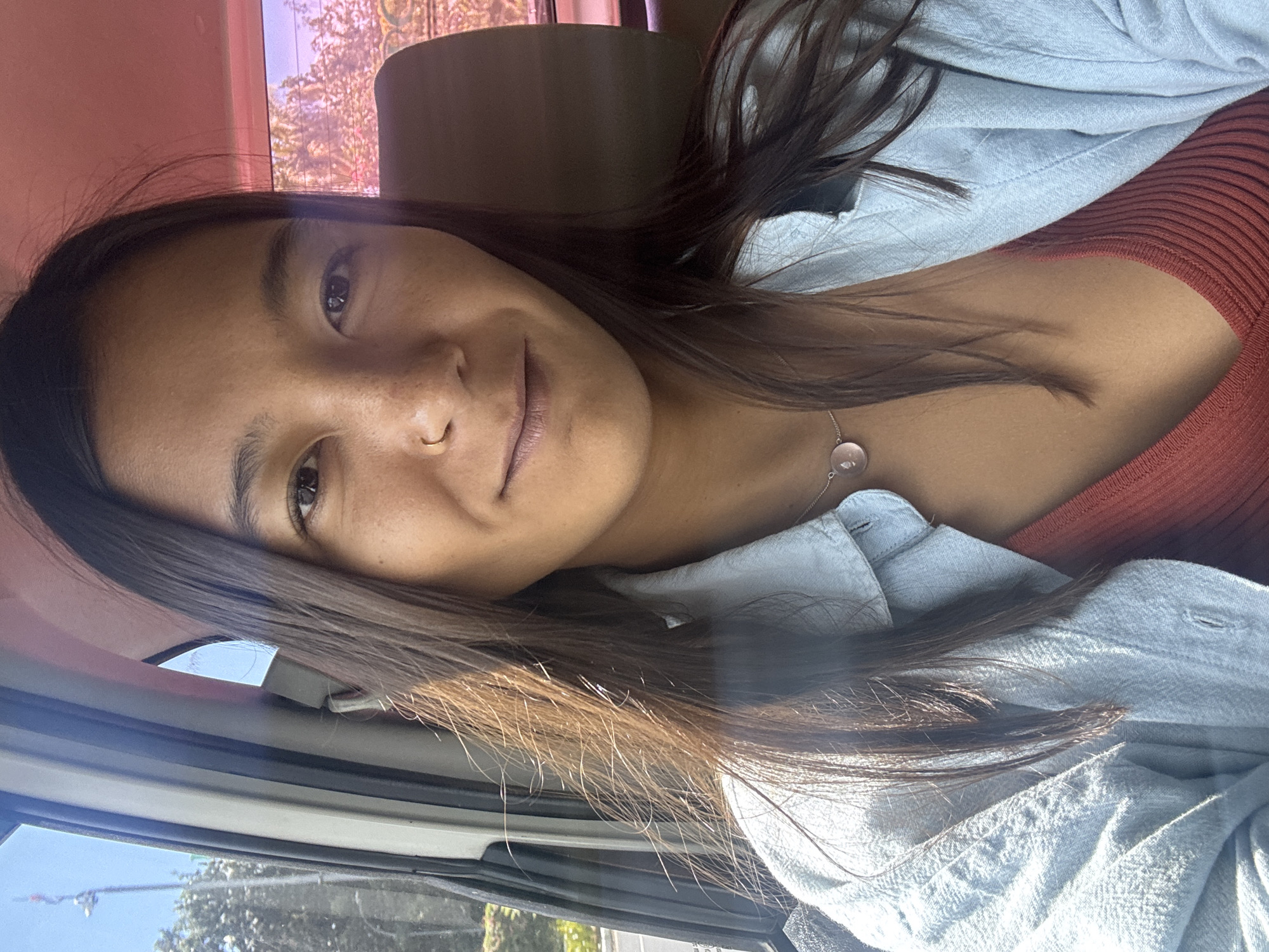 A young woman with long, dark brown hair and fair skin is taking a selfie from the front passenger seat of a vehicle. She is wearing a light blue button-down shirt over a rust-colored ribbed top and a delicate necklace with a round, pink pendant. A small gold hoop earring is visible in her left nostril. The background shows a blurred view of trees and a pinkish sky, suggesting it is either dawn or dusk, or a heavily filtered environment. The interior of the car is visible with a light-colored headliner and a dark-colored seat. The woman's expression is relaxed and she is looking directly at the camera. The lighting suggests a sunny day, with light rays illuminating her hair. The scene is consistent with being in Kuta, Indonesia, known for its vibrant coastal atmosphere and warm climate.