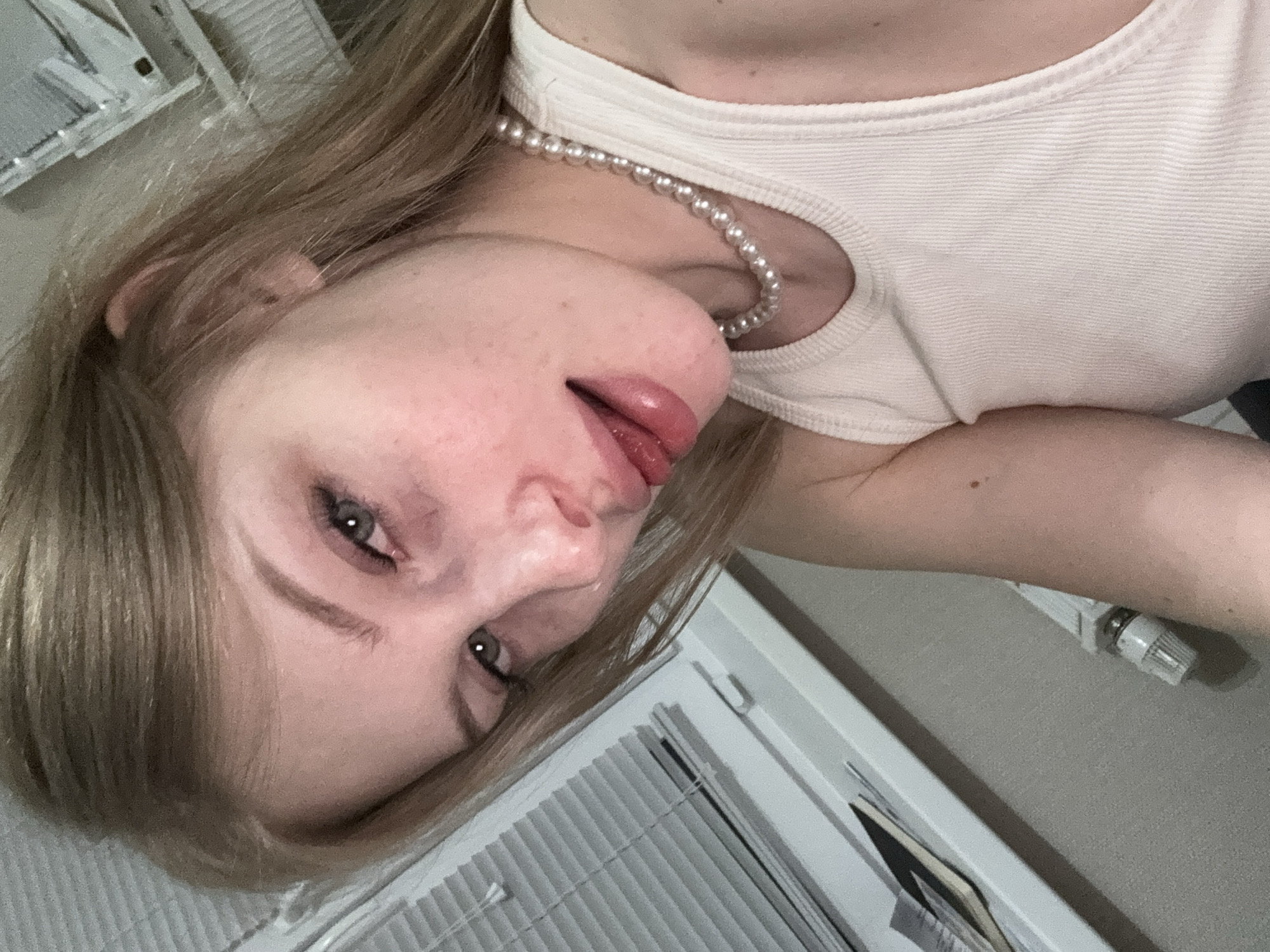 A young woman with blonde hair is captured in a close-up, slightly angled selfie. She is wearing a white tank top and a pearl necklace. Her gaze is directed towards the camera, with her head tilted and her tongue slightly protruding from her lips. The background shows blinds and a window, suggesting an indoor setting. The lighting is soft, creating a somewhat intimate atmosphere. There are no overt signs of specific location markers for Perm, Russia.