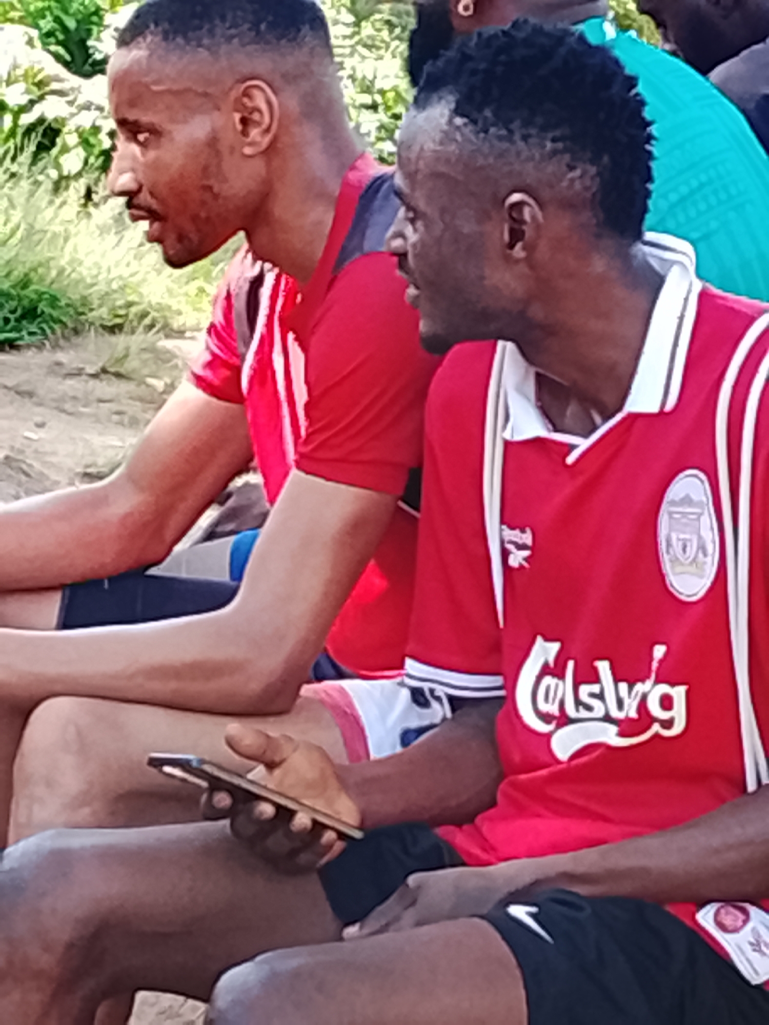 The image depicts two men sitting outdoors, likely in Jos, Nigeria, on a day with abundant natural light. The man on the left is wearing a red jersey with white stripes. The man on the right is also wearing a red jersey with white trim and a collar, featuring a crest on the left chest and the "Carlsberg" logo across the front. He is holding a mobile phone in his right hand. Both men appear to be of African descent and are dressed casually, suggesting a relaxed setting, possibly during a sporting event or a break. The background shows greenery, indicating an outdoor environment, possibly a park or a field.