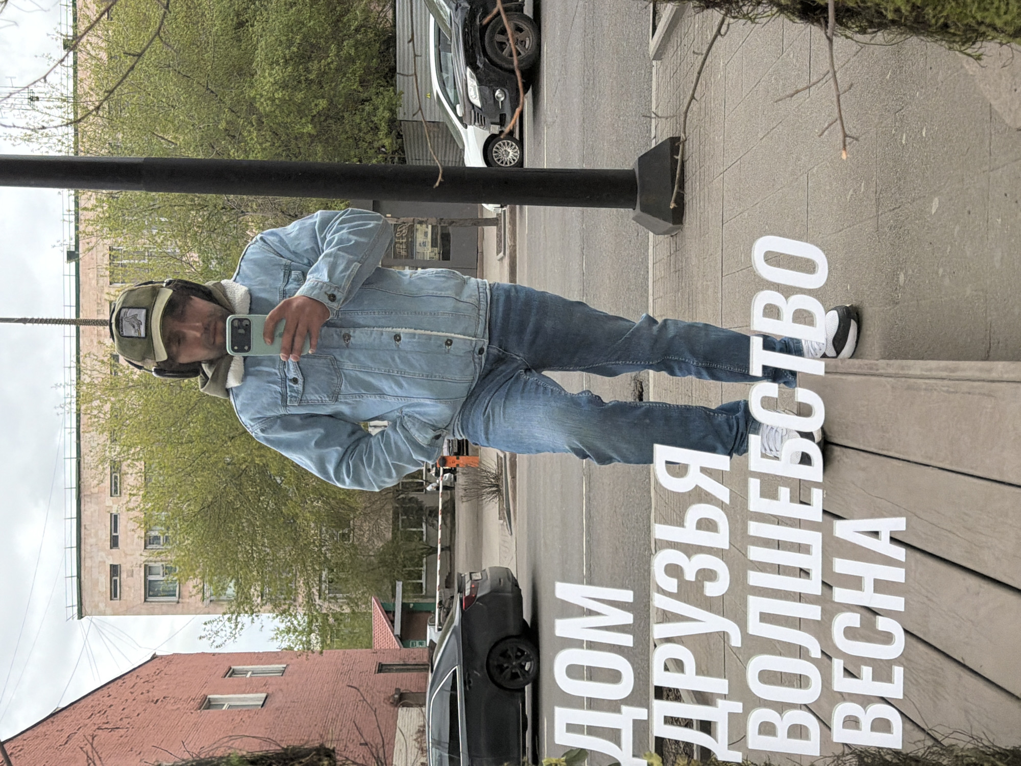 A man in a light blue denim jacket, jeans, and a cap with ear flaps is taking a selfie in front of a reflective surface. The reflection shows the environment around him, including a street with cars, buildings, and trees. In front of the reflective surface, there are large white letters that spell out a phrase in Russian. The letters seem to be part of an advertisement or sign. Based on the Russian script in the image, this is likely taken in Krasnoyarsk, Russia.