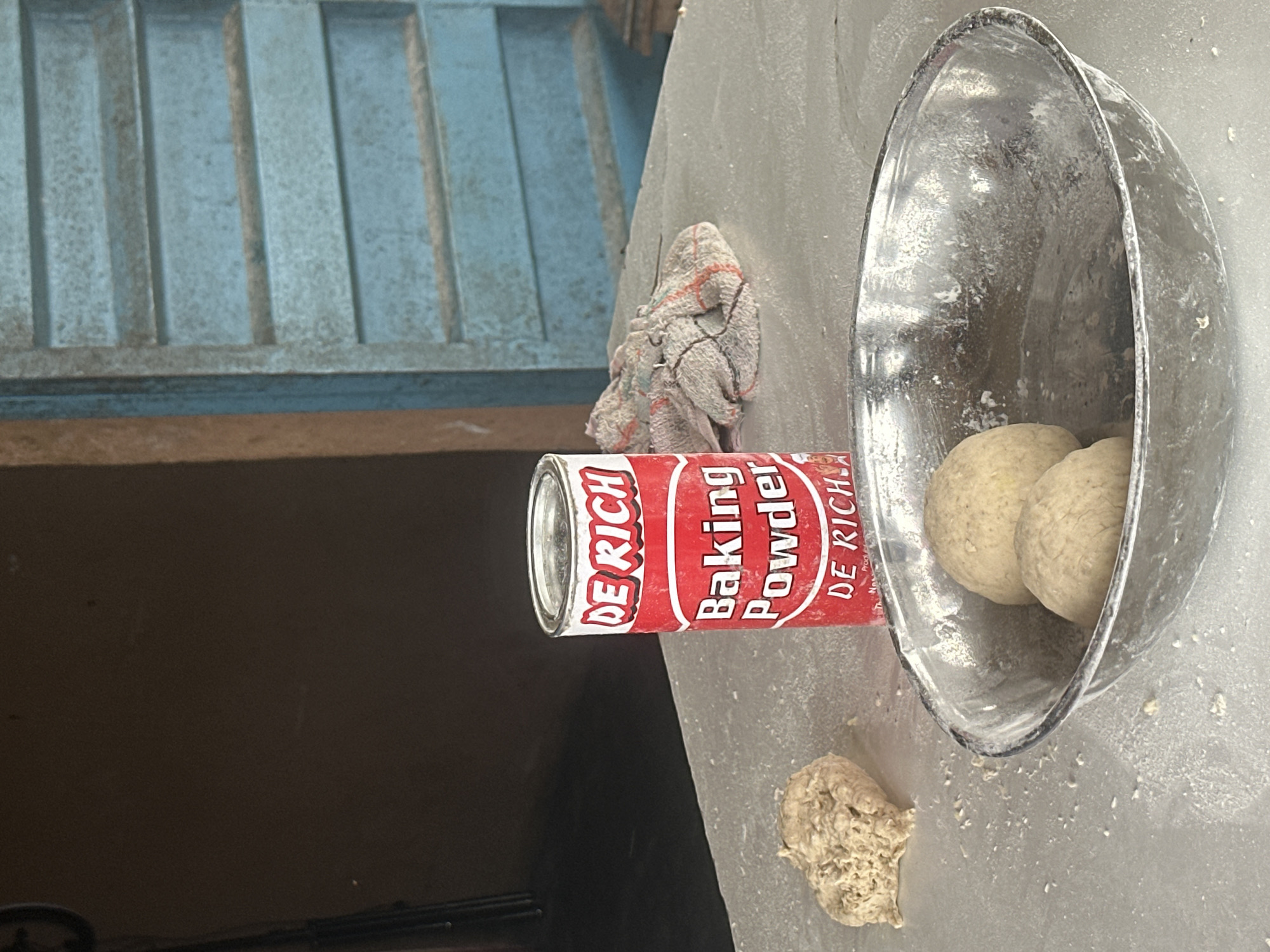The image depicts a close-up, eye-level shot of a table in what appears to be a home or small commercial space in Badagry, Nigeria. The table is covered in a fine dusting of flour. On the table, a red and white can of "DE RICH Baking Powder" stands upright. To the left of the can, a lump of dough sits, and in front of the can, a metal bowl contains two spherical portions of dough, also lightly dusted with flour. Behind the baking powder can, a colorful, patterned cloth is crumpled. The background shows a brown wall on the left and a light blue, slatted door or window on the right. The overall impression is a scene related to food preparation.