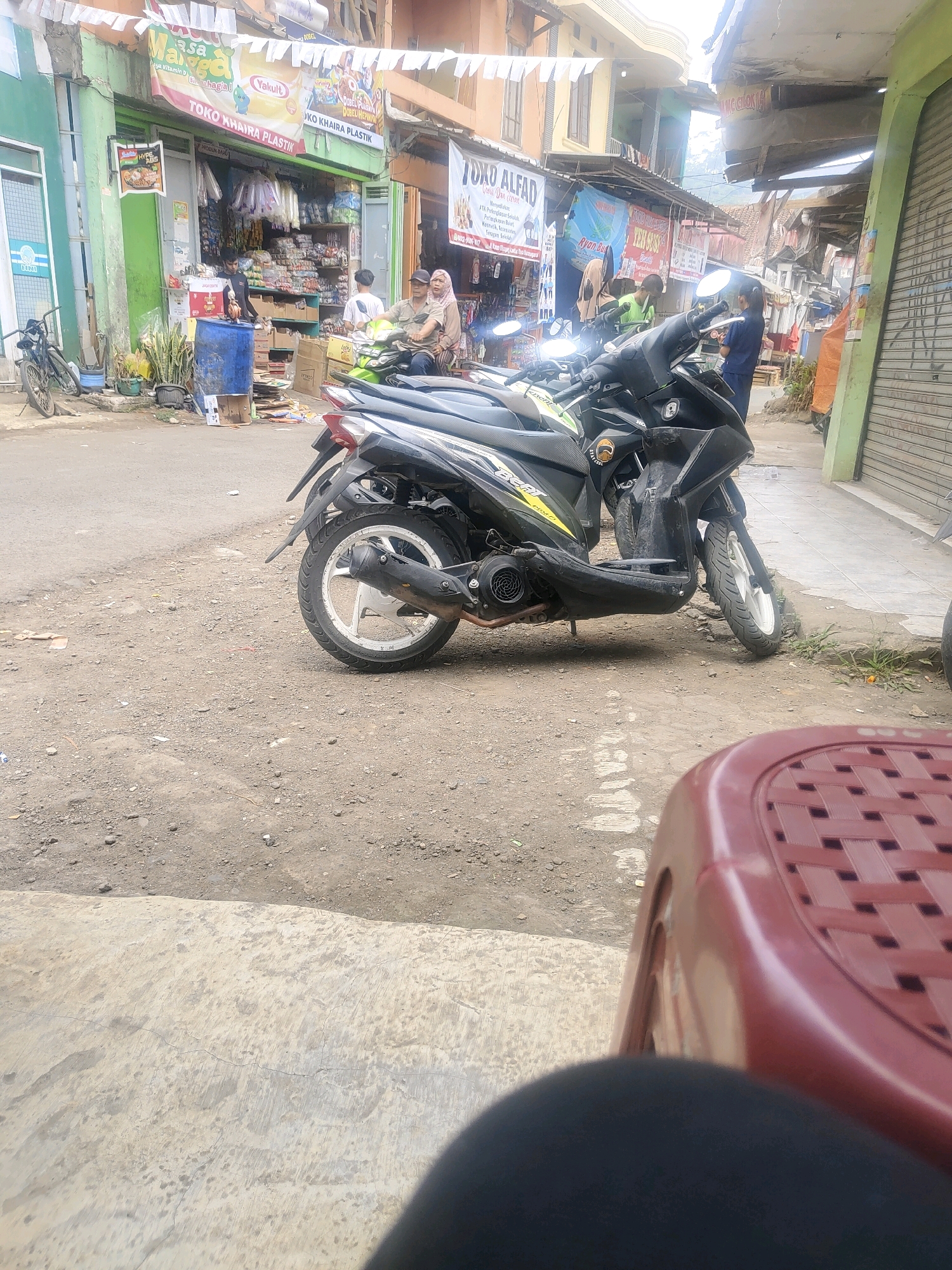 In Sukanagara, Indonesia, a street scene unfolds with several motorbikes parked near shops. A black scooter with yellow accents is prominently featured in the foreground, resting on a dirt and gravel road. To the left, a bicycle leans against a green-walled shop. Behind the parked motorbikes, two people are visible, one standing and one seated on a motorbike. Further back, a man stands in the doorway of a shop filled with merchandise. The shop, identified by signs including "TOKO KHAIRA PLASTIK" and "TOKO ALFA," appears to be a retail establishment. The background reveals more shops and buildings lining the street, suggesting a commercial area. The overall environment is that of a typical Indonesian street with a mix of businesses and parked vehicles.