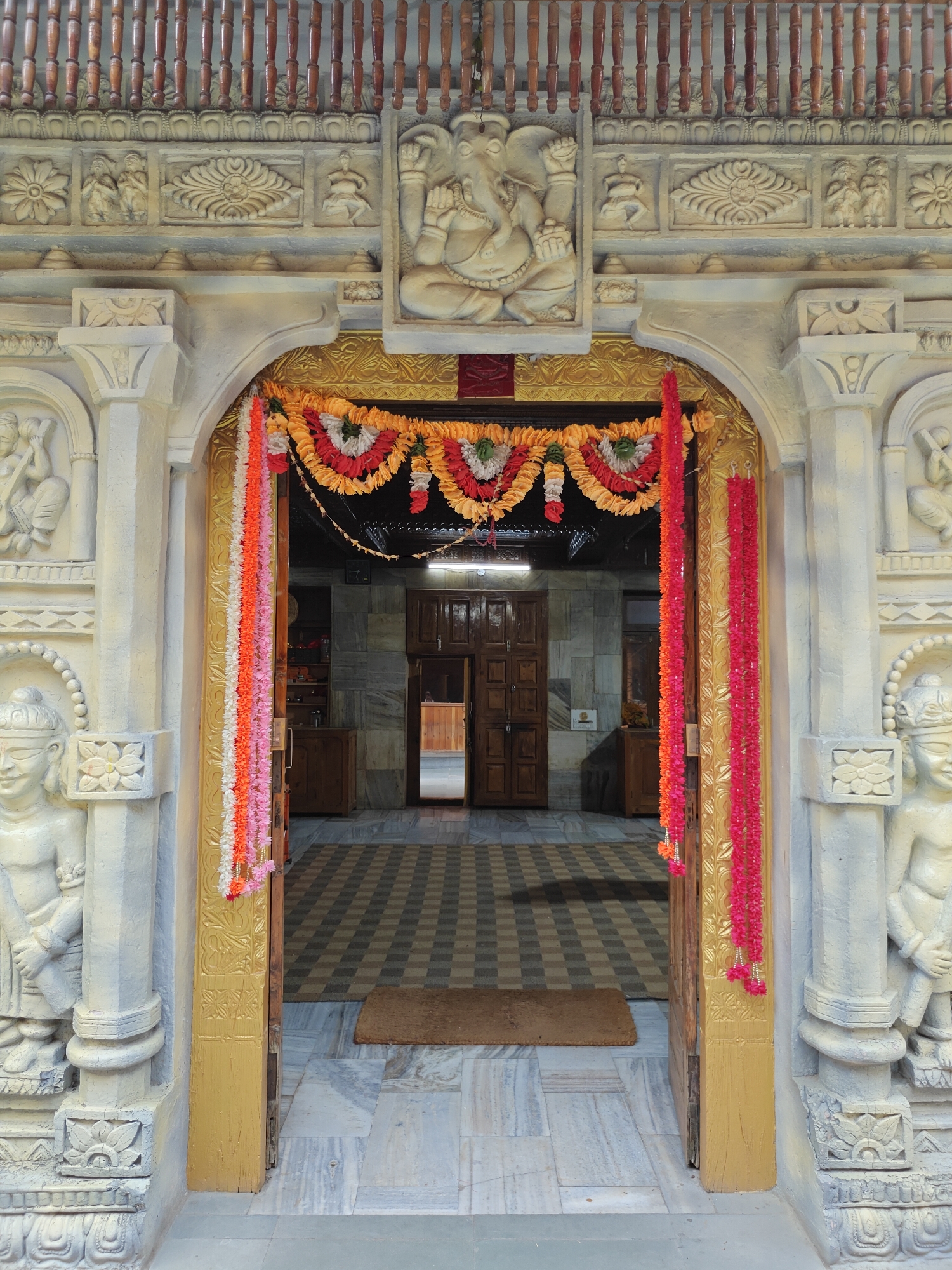 The image captures the intricately decorated entrance to a temple or religious building in Manikaran, India. The main subject is a grand archway, sculpted from a light-colored material, likely stone or plaster, featuring extensive carvings.
Above the archway, a prominent relief sculpture of Lord Ganesha, seated, is centered. Flanking Ganesha are panels depicting floral motifs and smaller human or divine figures in various poses, some seemingly devotional or performing actions. Above these carvings, a wooden balustrade with multiple turned spindles extends across the top of the frame.
The archway itself is framed by thick pillars on either side, each adorned with carved standing figures of deities or guardians, as well as decorative patterns and floral designs at their bases. The interior of the archway is painted gold, and draped across its upper opening are festive garlands made of artificial flowers in shades of red, orange, yellow, white, and pink. Additional vertical garlands hang down the sides of the golden door frame.
Beyond the open golden-colored wooden doors, a tiled floor with a black and white checkered pattern is visible, leading further into the building. A small, rectangular brown doormat rests just inside the threshold. The interior also features marble or tiled walls and wooden furniture, including what appears to be a cabinet and another closed wooden door in the distance.
The lighting suggests it is daytime, casting no harsh shadows. While no specific event is taking place, the colorful garlands indicate a celebratory or devotional atmosphere. There is no visible text in the image.