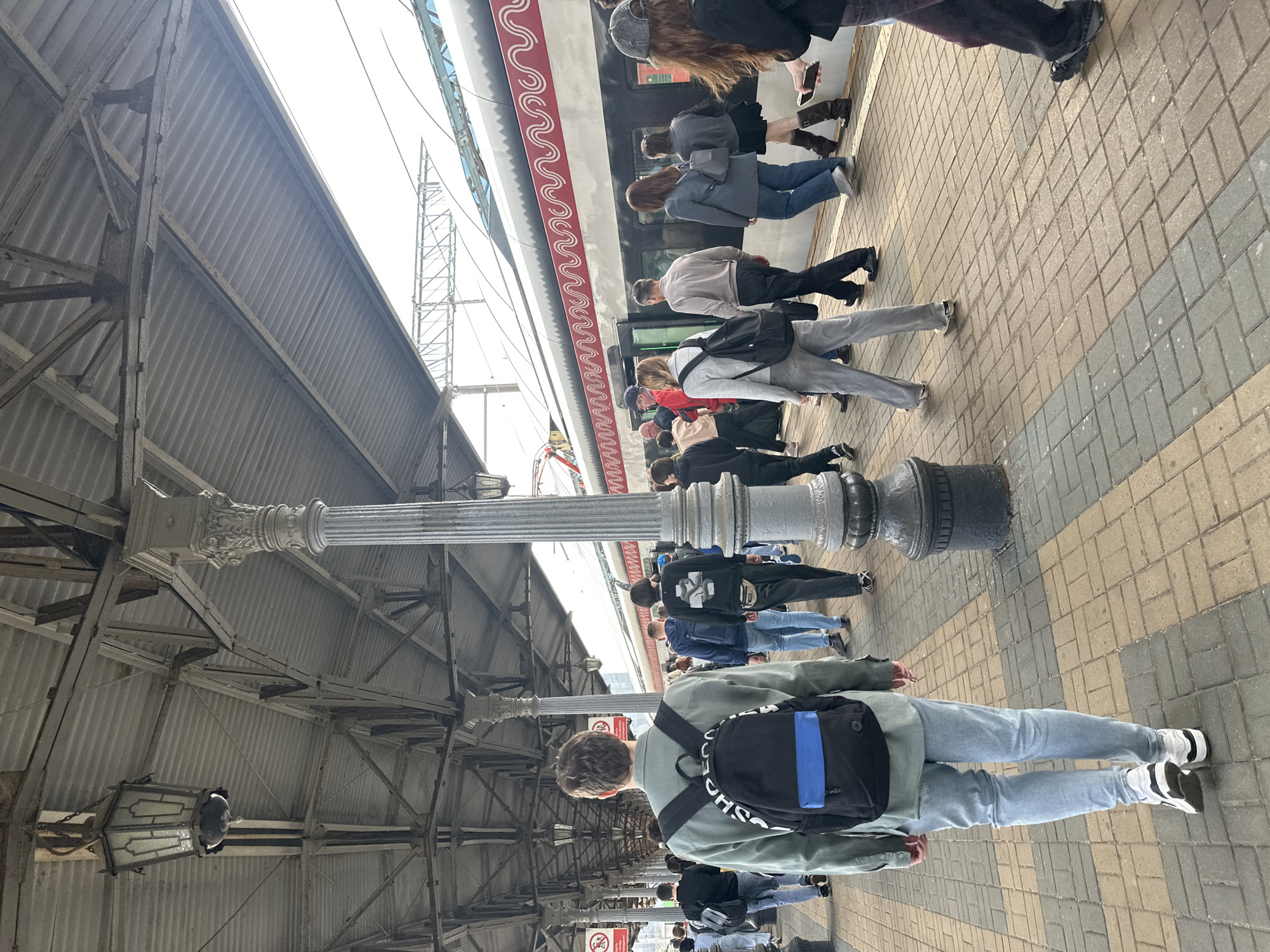 A group of people walk along a train platform in a city. The platform is covered by a metal roof supported by columns. A train is parked alongside the platform, and the people are walking towards or away from it. The ground is paved with rectangular bricks. The scene appears to be during the daytime, with natural light illuminating the area.