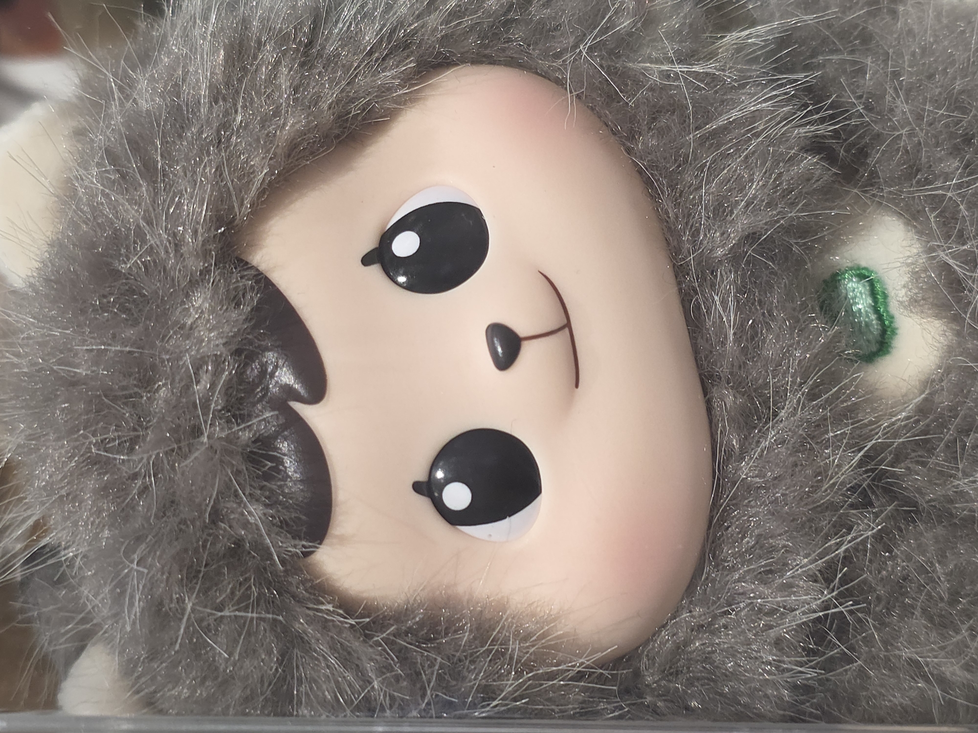 A close-up shot depicts a plush toy with a round, plastic face. The face features large, black eyes with white highlights, a small, triangular nose, and a curved, stitched smile. The cheeks are tinted pink. The face is surrounded by grey, shaggy fur. A small, green embellishment is visible near the edge of the fur on the right side of the frame. The setting or location within Warsaw, Poland, is indiscernible from this image.