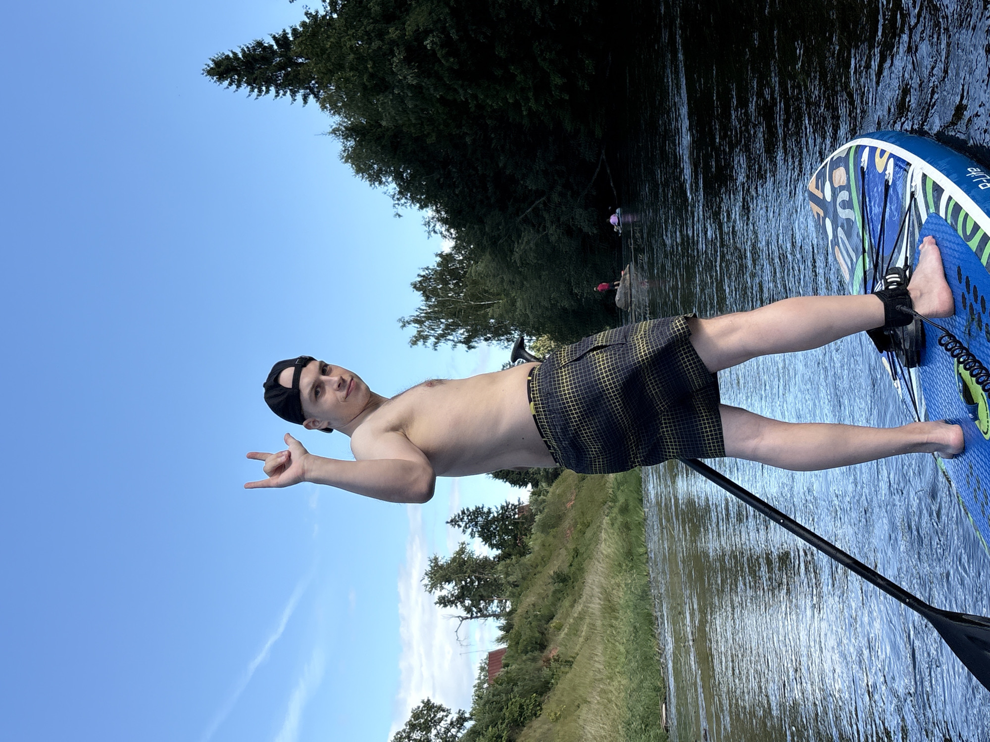 A young man with a shirtless torso and a black baseball cap stands on a blue paddleboard in a wide, calm river. He wears plaid swim trunks and has his right arm raised, making a rock-and-roll hand gesture. The paddleboard is equipped with a leash attached to his ankle. The river flows through a lush, green landscape with a gently sloping embankment on the left, covered in grass and trees, with a few buildings visible in the distance. The right bank is densely forested with tall trees. The sky is clear blue with a few wispy clouds. In the background, a few other people are visible on the water, some on kayaks or paddleboards. The scene appears to be set in the city of Siversky, Russia, likely during a warm, sunny day.