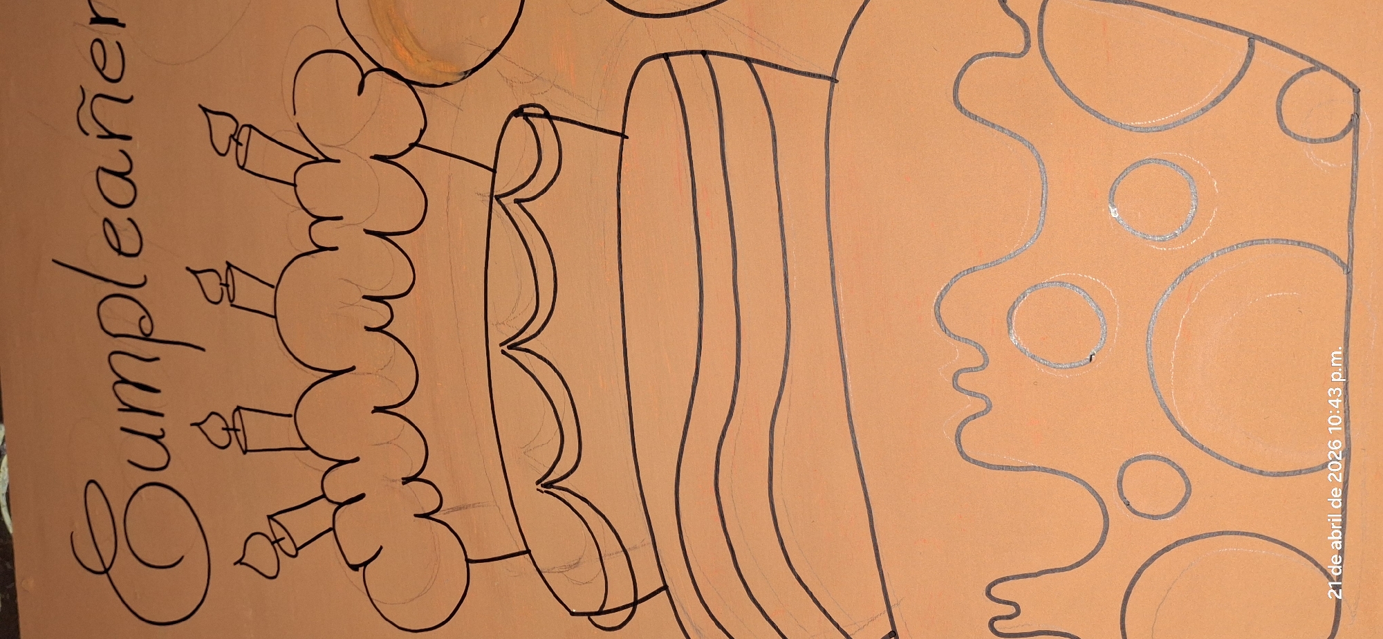 This media file captures a drawing of a birthday theme on a light orange-peach background, viewed in a portrait orientation but originally drawn in landscape. The primary subject is a large, multi-tiered birthday cake outlined in black, featuring what appears to be decorative icing or frosting along its layers and at the top. Several candles with flames are depicted on top of the cake, also outlined in black. To the left of the cake, the word "Cumpleaños" (Spanish for birthday) is written in elegant black script.
On the right side of the image, separated from the cake, are outlines of shapes that resemble a slice of cheese with circular holes or abstract organic forms, drawn with a lighter, possibly silver or light gray, outline. Faint pencil marks and sketches are visible underneath some of the black outlines, suggesting the drawing process.
The scene depicts an artistic creation, likely a drawing or painting in progress or recently completed, intended for a birthday celebration. The time of day indicated by the visible watermark is 10:43 p.m. on April 21, 2026. The location context for this media file is Vallecito, Venezuela.