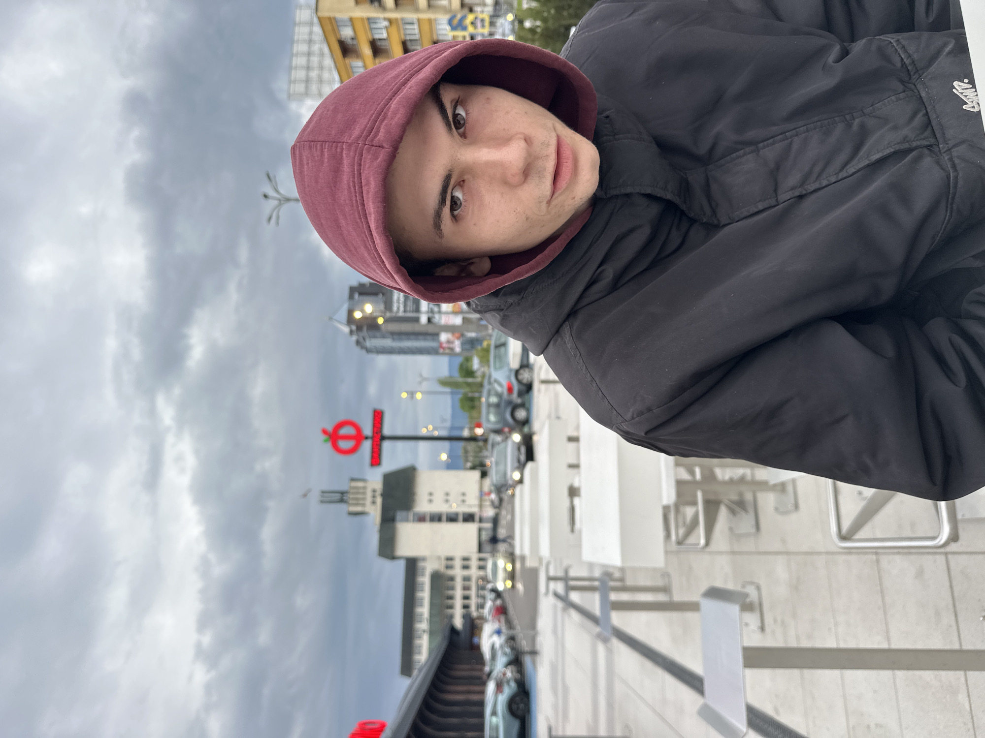 A young man is the primary subject of this outdoor portrait, positioned in the foreground and looking directly at the camera with a neutral expression. He wears a maroon hooded sweatshirt under a black puffer jacket, with the hood pulled up. His head is slightly tilted, and he has short dark hair with light facial stubble.
The setting is an urban environment under an overcast, cloudy sky, suggesting daytime without direct sunlight. In the background, several buildings of varying architectural styles are visible, alongside a street with parked and moving cars. On the right side of the image, white and silver metal structures, possibly outdoor furniture or barriers, create a dynamic foreground element.
A notable feature in the mid-ground is a distinctive red circular sign with a white horizontal bar, reminiscent of the London Underground logo, but in red. Inside the white bar, Cyrillic text is visible, which appears to read "БППЛОВ". This sign acts as a key location cue for the city of Plovdiv, Bulgaria, where Cyrillic script is used. The overall scene depicts a typical city street environment.