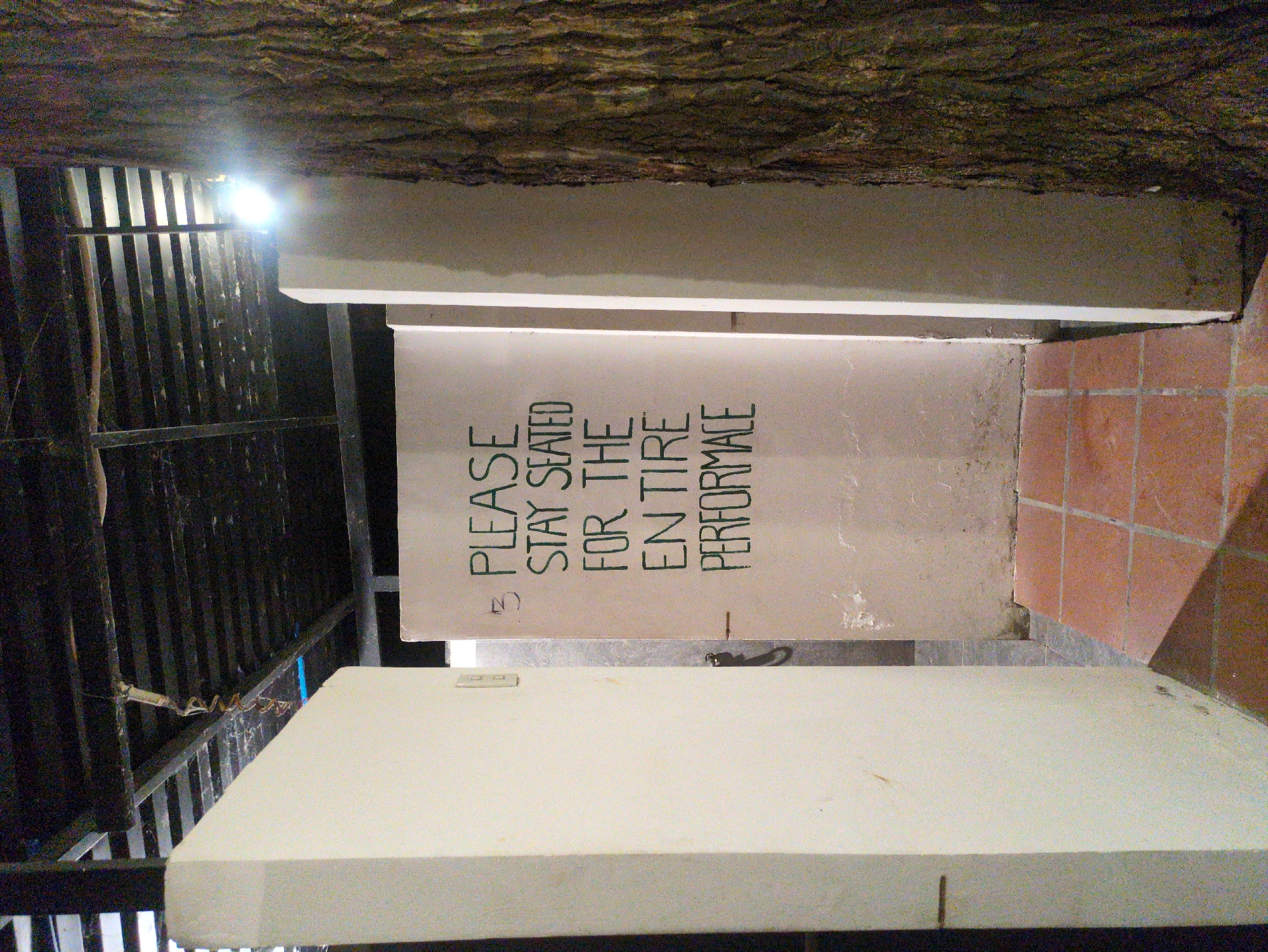 The image shows an indoor seating area with the message "PLEASE STAY SEATED FOR THE ENTIRE PERFORMANCE" displayed on a sign. The location is likely in Phú Quốc, Vietnam. The ceiling appears to be made of natural materials, possibly wood or bark. There are white concrete seating areas with metal railing nearby. The floor is tiled with brown square tiles. There are no people visible in the image.
