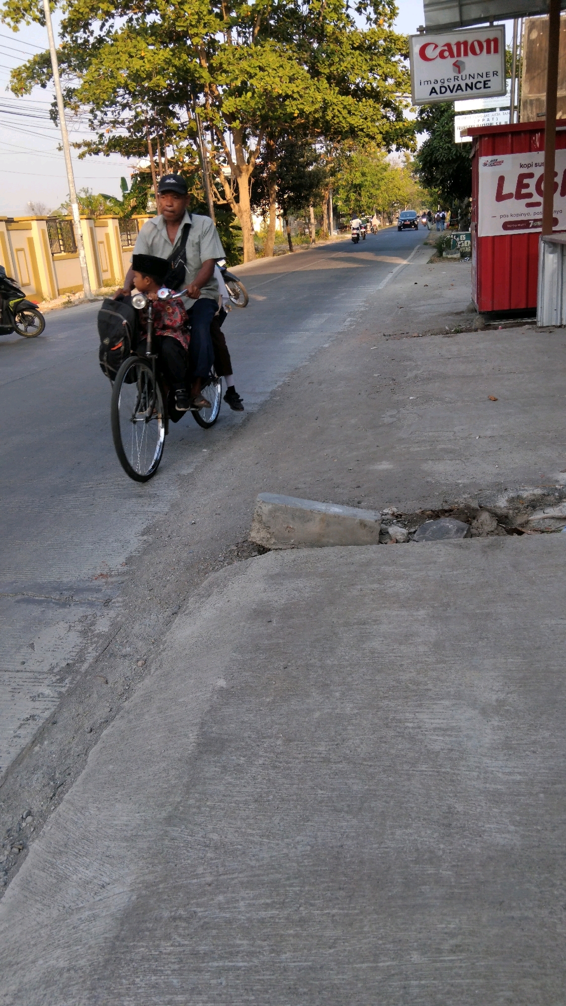 This is a vertical, medium-shot, eye-level photograph taken from a slightly elevated position looking down at a street scene in Tegal, Indonesia. In the foreground, a man is riding a bicycle with a child seated in front of him. The man is wearing a light-colored collared shirt, dark pants, and a black baseball cap. He has a dark bag slung across his chest. The child is wearing a dark cap and a patterned shirt. They are on a paved road with a concrete curb and sidewalk to their right. Further down the street, other people and vehicles, including motorcycles and a car, are visible. A red structure with a white sign that reads "LEGI" is on the right side of the street, along with a Canon imageRUNNER ADVANCE sign. Tall trees with lush green foliage line the street. The lighting suggests it is late afternoon or early evening, with the sun casting a warm glow.