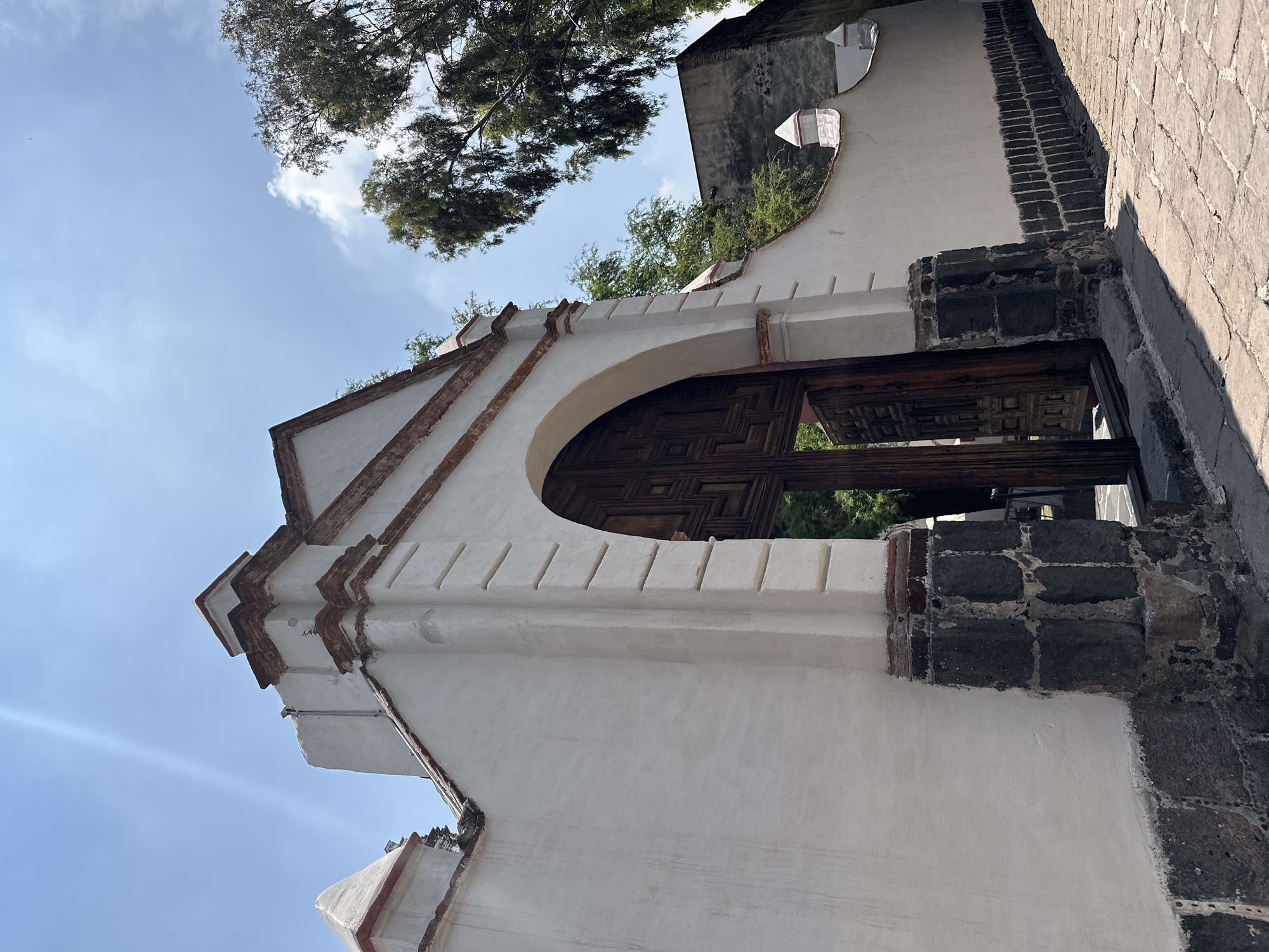 The image presents an upward-angled view of a traditional architectural structure, likely a gateway or part of a building, under a bright blue sky with scattered white clouds. The scene is set in daylight, indicated by the clear visibility and subtle lens flare in the upper left.
The central subject is a robust archway constructed from white stucco or plaster. Its design features decorative accents of reddish-brown brick or similar material along the edges and top, providing a striking contrast to the clean white surface. The base of the archway's right support is built from dark, rough-hewn stone blocks, possibly volcanic rock, a common building material in the region, which adds to the rustic charm.
Behind the archway, a large, dark wooden door or gate is visible, distinguished by intricate geometric carving patterns. The door appears to be slightly ajar or set back, revealing glimpses of vibrant green foliage and tree branches, suggesting a garden or interior courtyard beyond.
To the immediate right of the archway, a paved pathway or street made of irregular stone blocks or cobblestones extends into the background, hinting at an older urban or historical setting. Overhead, leafy green trees frame the sky, adding an organic element to the architectural focus. The image conveys a sense of historical character and natural integration, consistent with the given location context of Mexico City. No discernible text is visible.