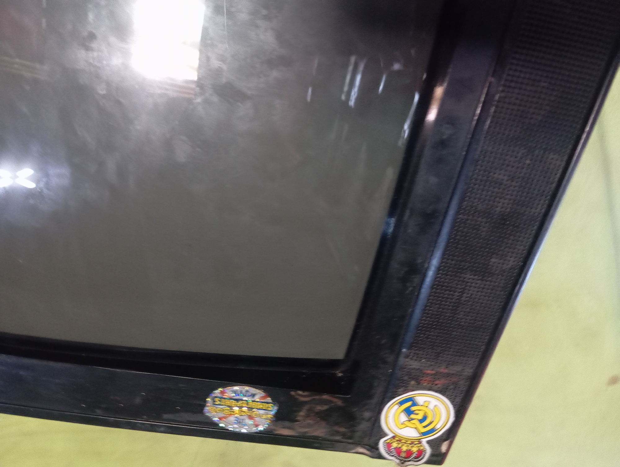 The image shows a close-up of the top-left corner of an older, black television set. The television has a textured speaker grille on the left side and a black bezel around the screen. Affixed to the top bezel are two stickers. The sticker on the left is round and features a crest with a crown on top, a shield with blue and yellow elements, and the letters "R.M." within the shield. This is the crest of Real Madrid Football Club. To the right of the Real Madrid crest is another round sticker with a holographic, iridescent design that appears to be a promotional or certification sticker, but its details are largely obscured by the reflective surface of the TV. The background is a plain, light green wall with some smudges. There are no people or actions depicted. The scene is consistent with an indoor setting. The image does not provide any information about the location in the city of Rejosari, Indonesia.