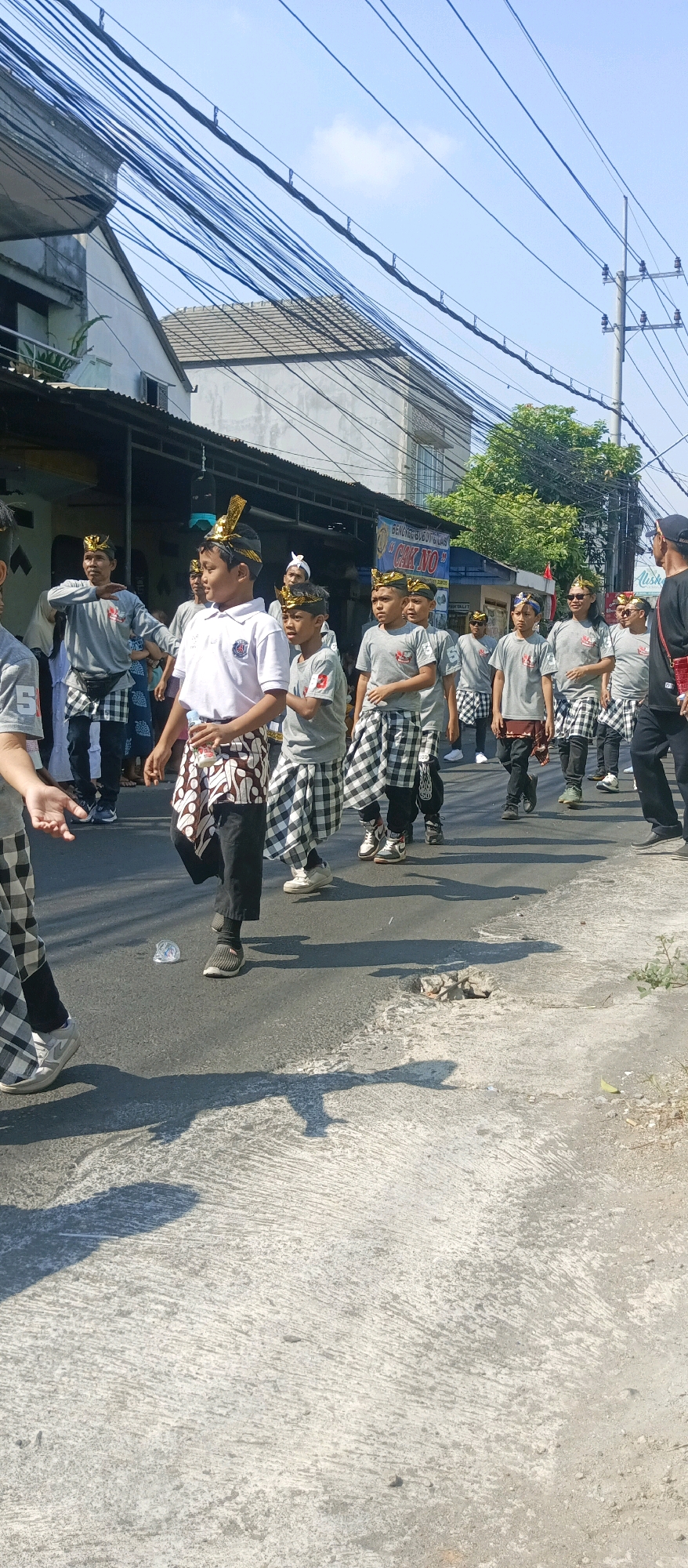 A group of young boys in traditional Balinese attire are participating in a parade on a street in Malang, Indonesia. They are wearing grey t-shirts, black pants, and black and white checkered sarongs. Some of the boys have on gold headpieces. They are walking in a procession. A few adults are present, also in traditional attire. The street is paved and lined with buildings and trees. The sky is clear and blue. The image captures a cultural event.
