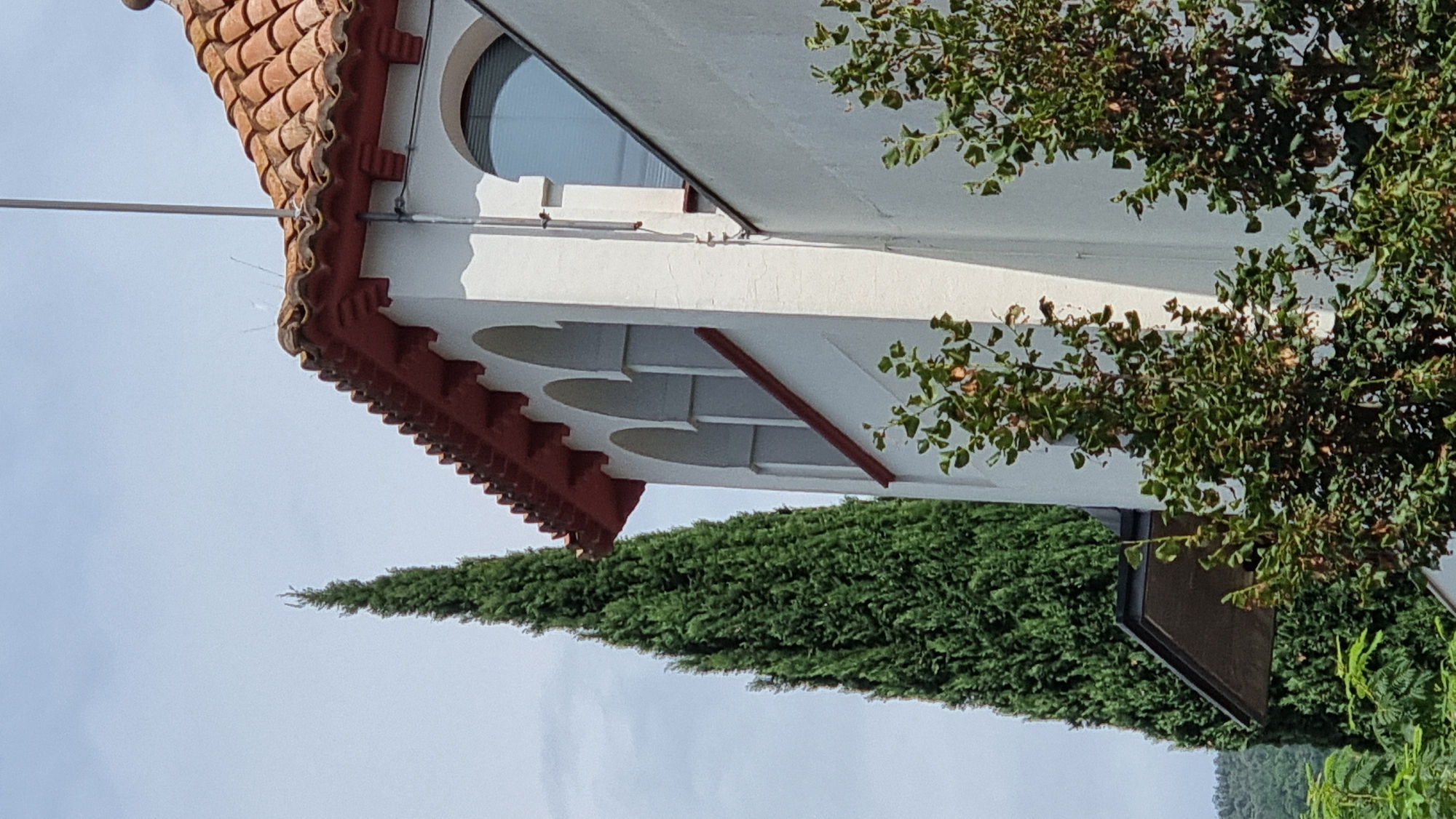 A white building with a red-tiled roof and arched windows is shown against a cloudy sky in Sant Joan les Fonts, Spain. A tall, green evergreen tree stands to the left of the building, and smaller green bushes are in the foreground. The building appears to be a bell tower or a similar structure with decorative red trim. The sunlight suggests it is daytime.
