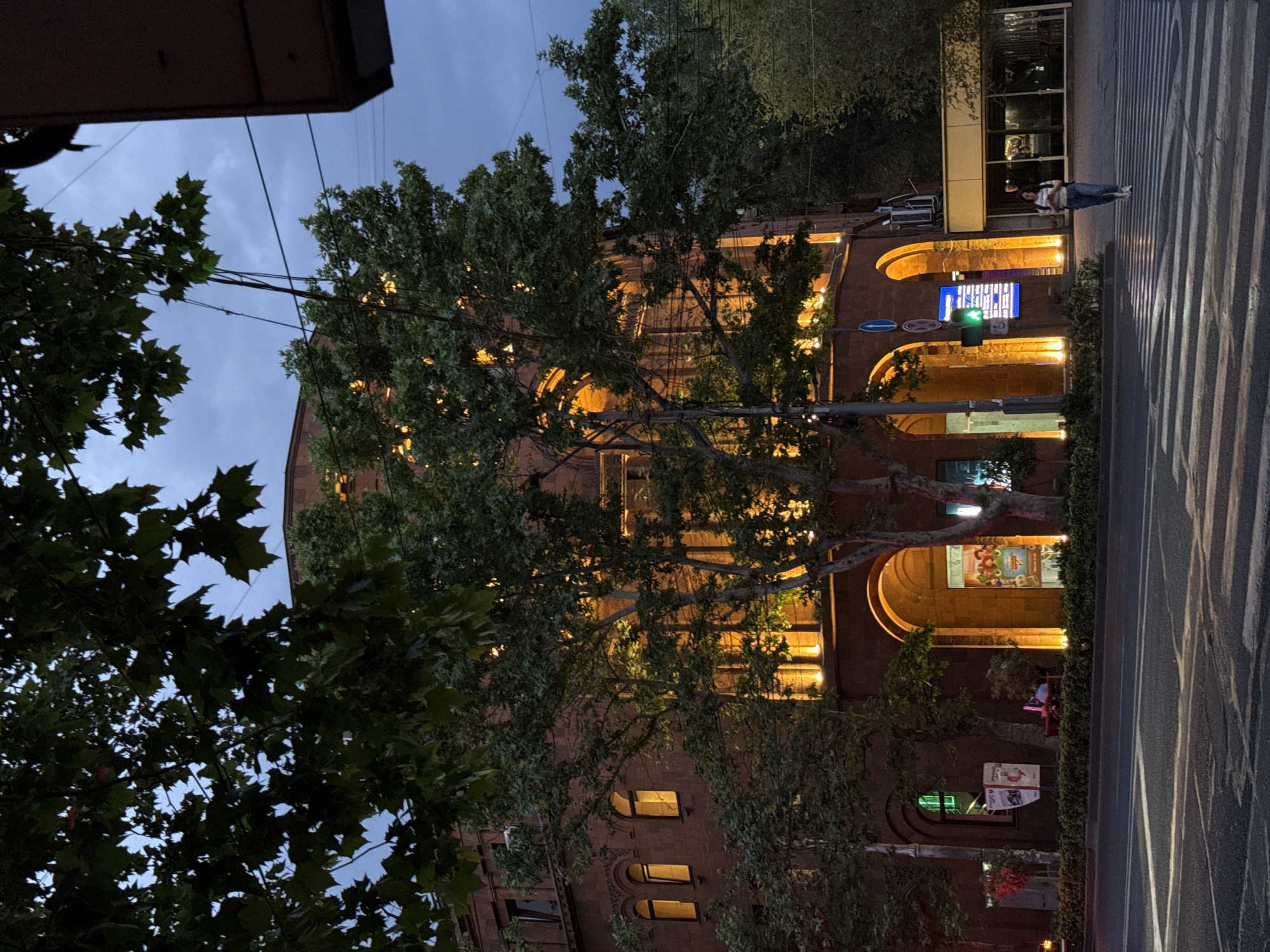 This image captures a vibrant urban street scene in Yerevan, Armenia, at dusk or early night. The central focus is a multi-story building, likely constructed from reddish stone or brick, with a distinctive architectural style featuring multiple arched windows and doorways. The building's facade is warmly illuminated by yellow lights, highlighting its details and creating an inviting glow that filters through the surrounding trees.
Lush green trees with dense foliage are prominent, positioned both in the foreground and mid-ground. Their branches partially obscure the building, adding depth to the scene while allowing glimpses of the illuminated architecture.
To the right, a paved street with a clearly marked zebra crossing (pedestrian crosswalk) is visible, its bright white stripes standing out against the dark asphalt. A solitary individual, dressed in light-colored clothing, is seen walking or standing near this crosswalk on the pavement. Further down the street, a small group of people appears to be seated at an outdoor establishment, suggested by red chairs.
The sky overhead is a dark blue-grey, indicating that it's either late dusk or early night. Artificial lighting is a dominant feature, including the building's warm facade lights, a brightly illuminated blue commercial sign mounted on the building (text illegible), and a vibrant green traffic light at an intersection. A street sign with a red circle and diagonal line (likely indicating "no stopping" or "no parking") is also visible. The scene conveys a lively urban atmosphere as evening sets in.