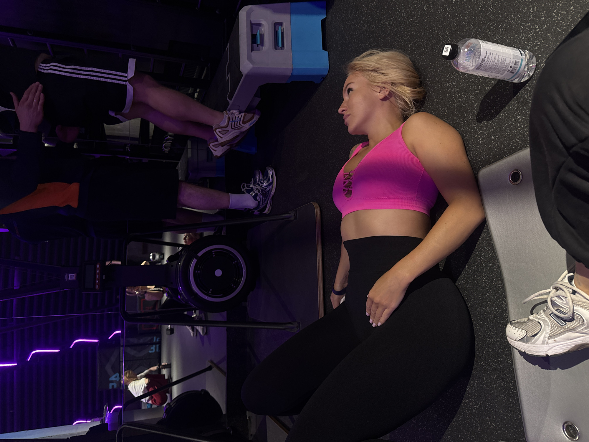 This image depicts a modern fitness gym, identified as being in Moscow, Russia, based on Cyrillic text visible on a water bottle.
In the immediate foreground, a blonde woman lies on her back on a dark grey speckled gym floor. She wears a vibrant hot pink sports bra and black leggings. Her hands are positioned on her upper thighs, and a blue fitness tracker is present on her right wrist. She looks upwards. To her right, a clear plastic water bottle with a black cap and a label predominantly featuring Russian text rests on the floor. Partially visible in the bottom left, a person's leg and foot, wearing a white and grey New Balance sneaker, are positioned on a grey mat.
In the mid-ground, a grey and blue adjustable step platform is present. A man wearing black shorts with white stripes and white sneakers stands on this platform, appearing to perform an exercise. A ring is visible on his left hand. To his left, another man is partially visible, wearing an orange and black top and black shorts.
The background features multiple pieces of gym equipment, including two rowing machines; the flywheel and monitor of one are clearly visible. The gym interior is dimly lit, with prominent purple LED strip lighting arranged in abstract patterns on the dark walls. In the far left background, another woman in a white top and red pants is seen bending down near a rowing machine. The scene suggests an active training session in a distinctly lit environment.