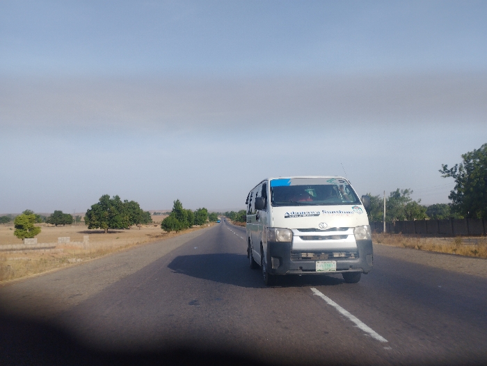 A white van with the words "Adanna Sunshine" in blue lettering is driving down a paved road in Kumo, Nigeria (10.067, 11.176). The van has a blue stripe on its side and a license plate. There is a white line down the center of the road. Trees are visible on either side of the road, with a light brown, grassy field in the background. The sky is a light blue with a faint, white haze.  The van appears to be traveling from left to right. The driver is not visible but there is a faint silhouette of another person in the passenger seat.  There is another vehicle visible in the distance, driving in the opposite direction, but it is too far away to be identified.