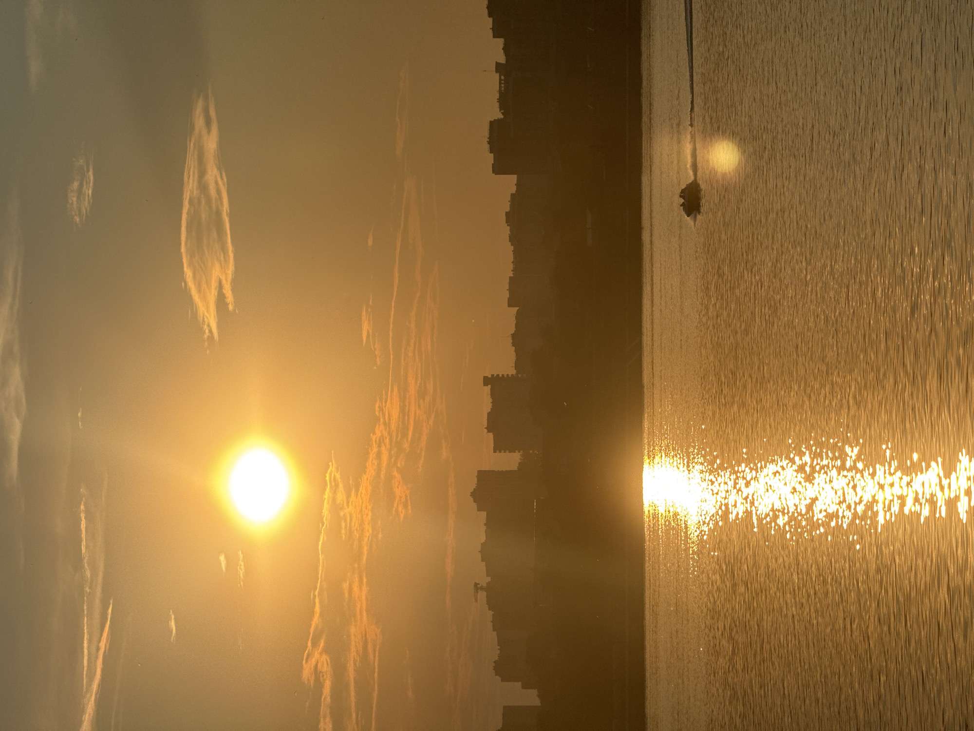 The image depicts a sunset over a body of water, likely the Angara River, in Irkutsk, Russia. The sun, a bright orb of golden light, is positioned in the upper left quadrant of the sky, casting a brilliant reflection on the water. Silhouetted against the warm glow of the sunset is a cityscape, with buildings of varying heights forming a distinct skyline along the horizon. In the foreground, the water is rippled, creating shimmering patterns of light. A small motorboat with at least two occupants is visible in the mid-right portion of the image, creating a wake as it moves across the water. The overall atmosphere is serene and picturesque, characterized by the warm hues of the sunset and the tranquil setting.