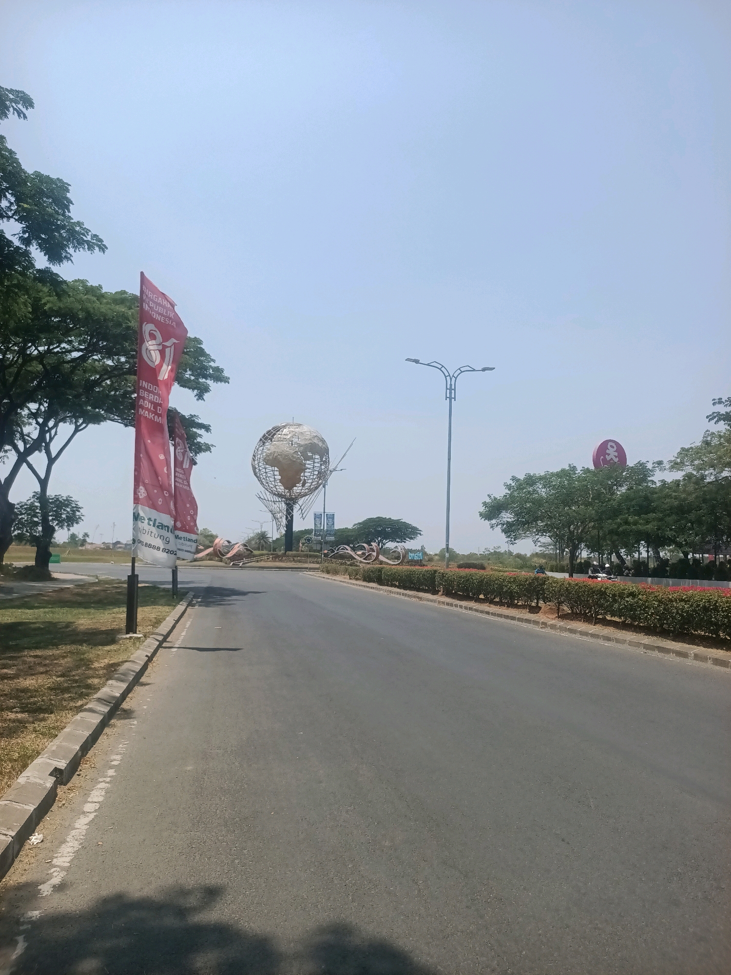 A wide, paved road stretches into the distance under a bright, clear sky. On the left side of the road, two red banners on poles display the number "81" in large white numerals. The banners also feature text in Indonesian, including "DIRGAHAMA" and "REPUBLIK INDONESIA," suggesting a celebration or commemoration. Beyond the banners, a cluster of trees with green foliage lines the roadside. Further down the road, a large globe sculpture with a metallic frame and a concrete earth element is visible. To the right of the globe, a modern streetlamp with multiple arms illuminates the area. In the background, more trees and a red circular sign with a "OK" logo are present. The road is bordered by a neatly trimmed green hedge and a sidewalk. The scene appears to be an entrance or a prominent landmark within the city of Telagamurni, Indonesia.