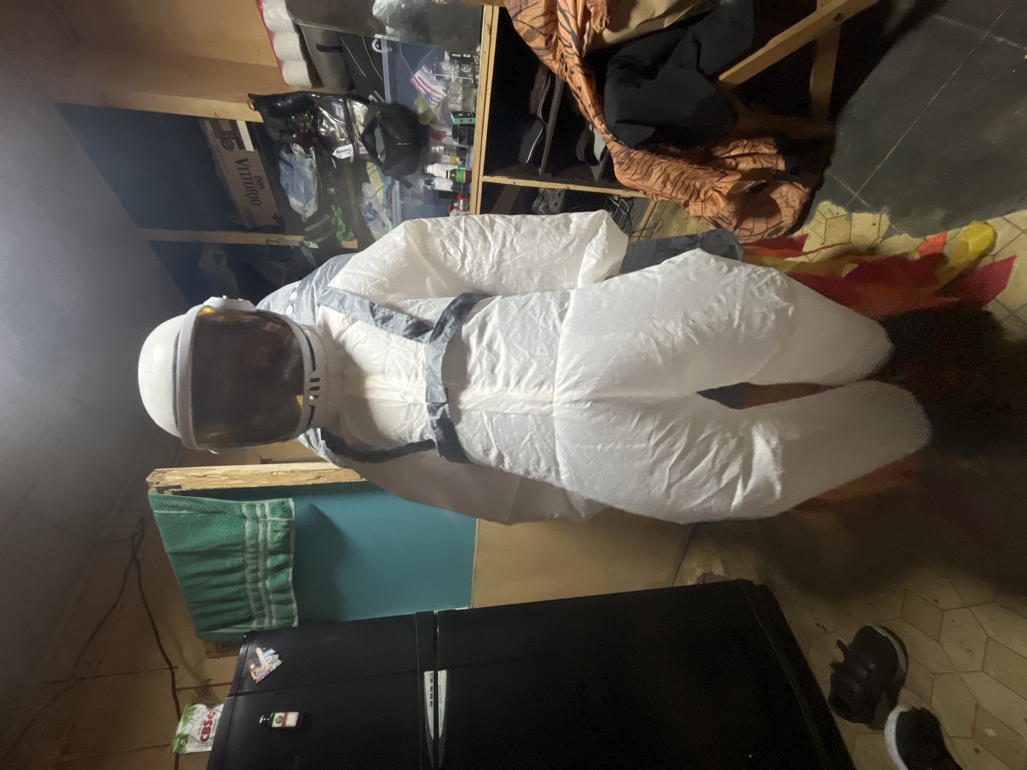 An indoor scene depicts an inflatable astronaut costume standing upright. The costume is primarily white with gray straps. It has a white helmet with a tinted visor. The background includes a black refrigerator, a green towel hanging on a wooden structure, shelves stocked with various items, and a wooden chair with fabric draped over it. The floor is tiled. The image appears to be taken in a residential setting, possibly a garage or storage area, not in the city of Iquique, Chile.
