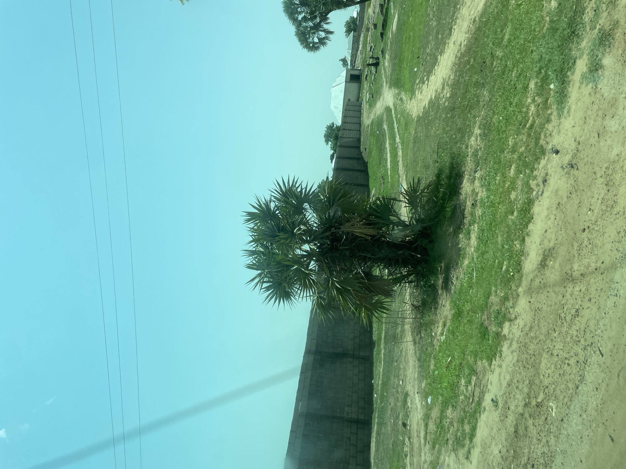 The image depicts an outdoor scene with a clear, pale blue sky above. In the mid-ground, a low, rough-hewn wall constructed from grey concrete blocks extends horizontally across the frame. In front of this wall, a dense palm tree with spiky fronds stands prominently. Beyond the wall, a path winds through a grassy area dotted with rocks. In the distance, several buildings with light-colored roofs are visible. The foreground consists of a dusty, unpaved surface with patches of grass and some scattered debris. A faint, diagonal shadow or reflection crosses the upper left portion of the image, possibly from a window or vehicle. The overall environment appears to be a rural or semi-rural area within Bauchi, Nigeria, characterized by its arid landscape and the presence of traditional building structures.