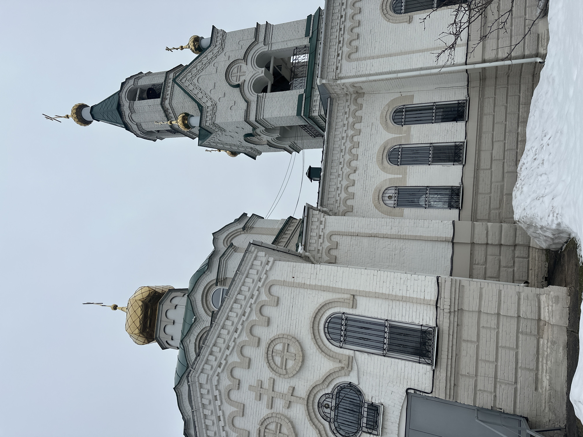 The image depicts a white brick Orthodox church located in Votkinsk, Russia. The architectural style features a combination of traditional Russian elements, including golden onion domes topped with Orthodox crosses, arched windows with decorative iron grates, and ornate brickwork patterns incorporating crosses. The church appears to be situated in an urban setting, with a patch of snow visible in the foreground. The sky is overcast, suggesting a winter or early spring day. There are no people visible in the image.