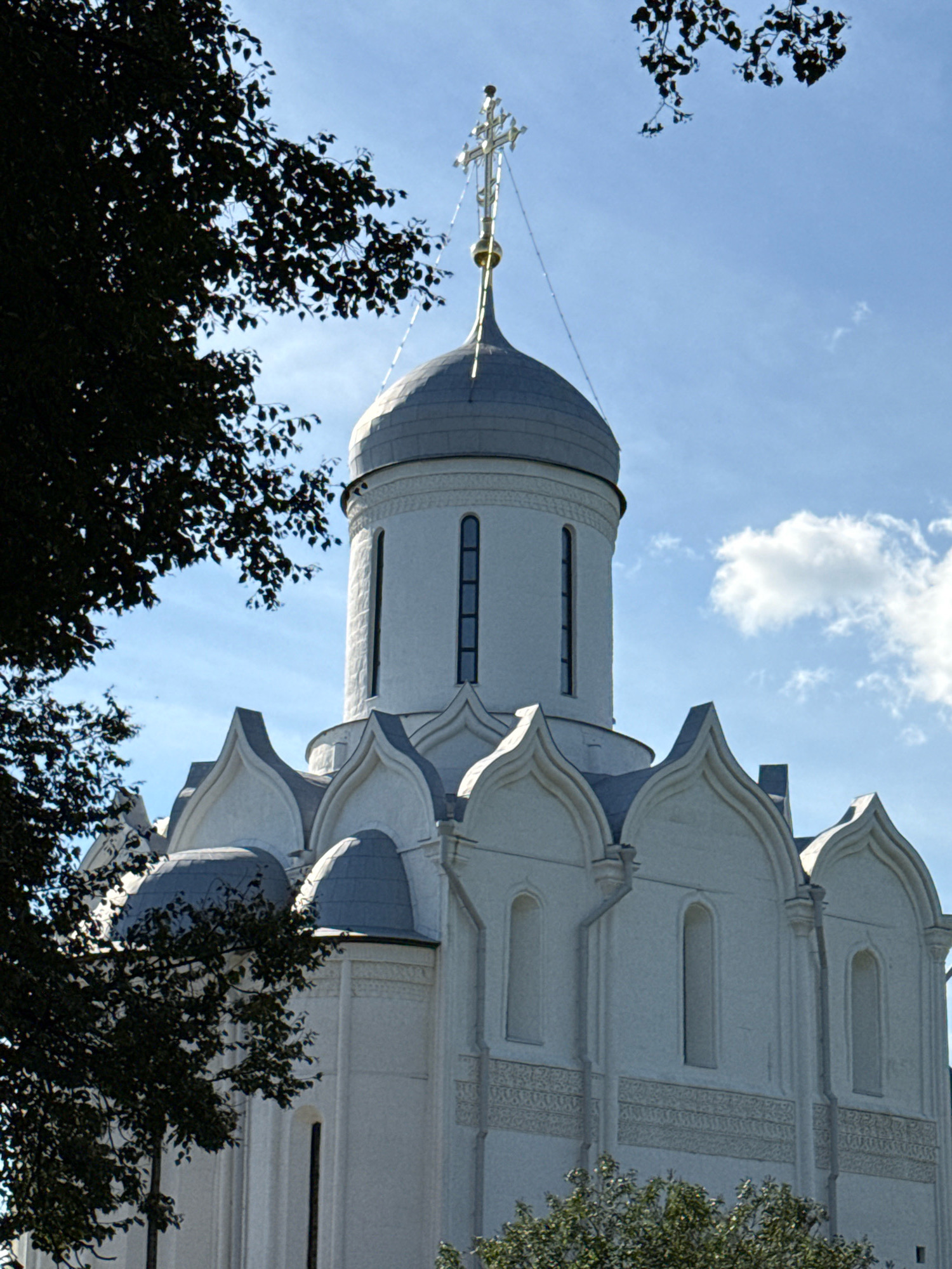 The image shows the Church of the Nativity of the Theotokos in the Gorodskoy District of Zvenigorod, Russia. The church is made of white stone and features a large central dome topped with a golden cross. The building has multiple smaller domes, arched windows, and intricate decorative carvings along its facade. Dark green foliage from nearby trees partially obscures the left side of the structure. The sky is blue with scattered white clouds, and the sun appears to be shining.