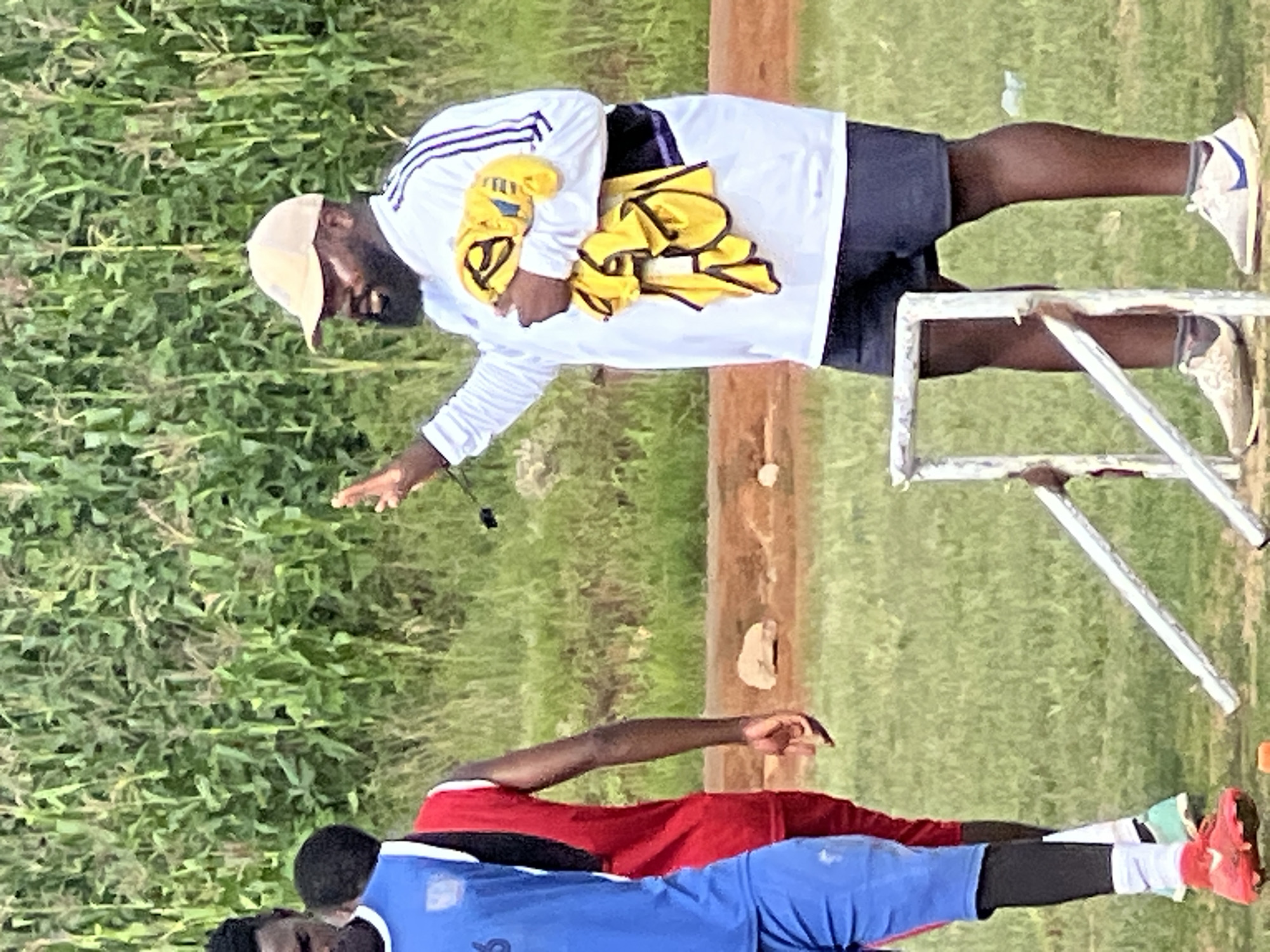 In Jos, Nigeria, a man wearing a light-colored baseball cap, a white long-sleeved shirt with dark stripes on the sleeves, and dark shorts stands outdoors. He is holding a yellow towel in his right arm and has his left hand raised, as if speaking. To his left, two young men are visible; one is wearing a blue jersey and blue shorts, and the other is wearing a red jersey and dark leggings. They appear to be on a grassy field with a dense green cornfield in the background. A metal hurdle frame is in the foreground. The lighting suggests it is daytime.