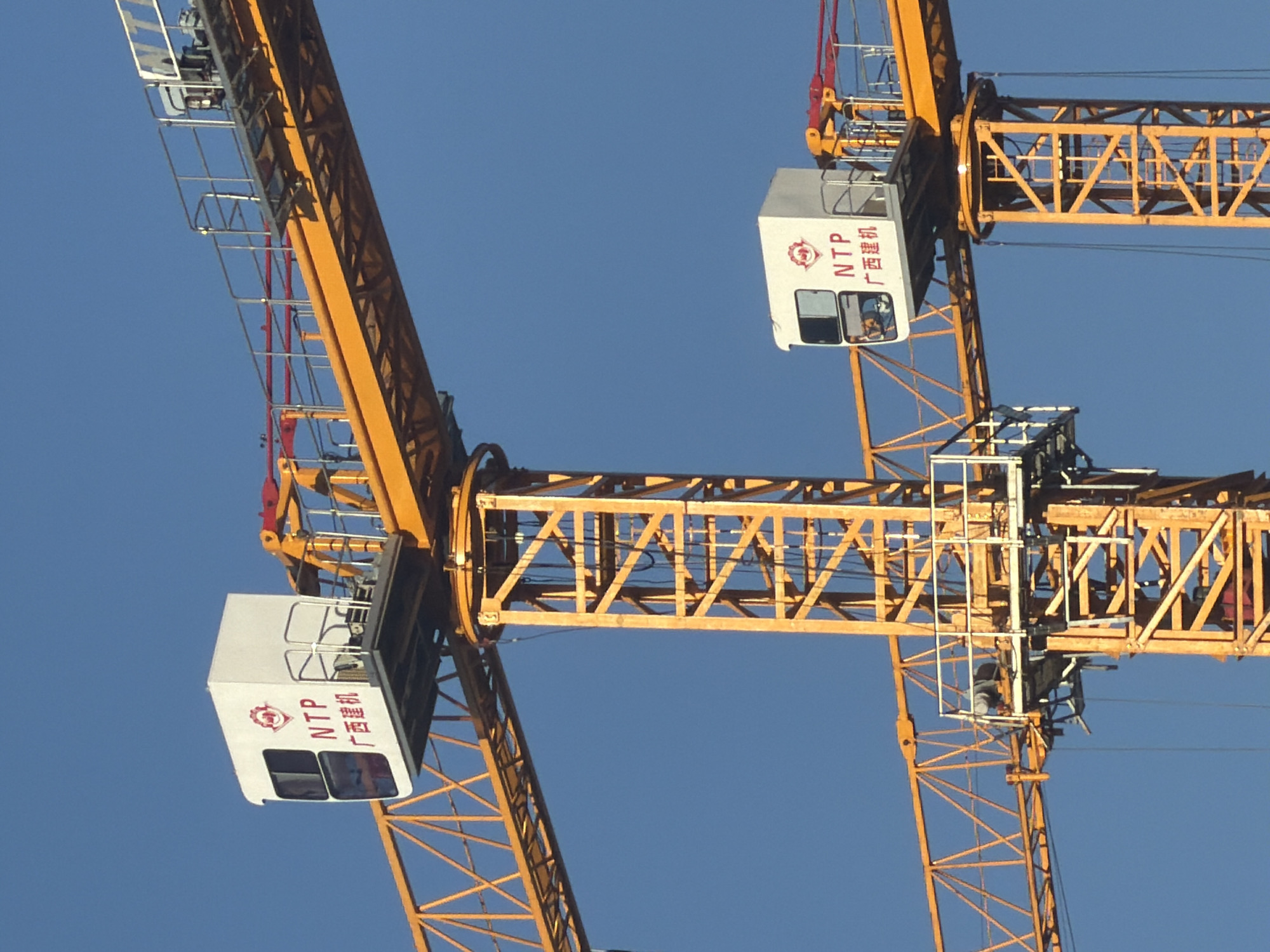 The image shows a close-up view of two yellow construction cranes against a clear blue sky. The cranes are positioned so that their central towers and horizontal jibs are visible. Both cranes have white operator cabs mounted on the towers, with the same red logo and text in both Chinese and English: "NTP 广西建机". On the upper crane's jib, there is a red metal safety railing. On the lower crane, parts of the tower structure and cabling are visible. The context suggests these cranes are part of a construction site. There are no discernible geographical markers in the image that would specifically place it in Astana, Kazakhstan.