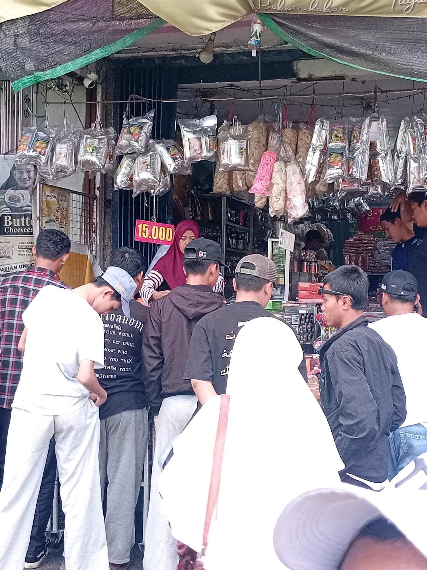 A group of people are gathered around a street stall in Lawang, Indonesia, likely purchasing snacks or goods. The stall is covered by an awning and displays various packaged items hanging from racks, including chips, crackers, and what appear to be food products. A price tag displaying "15.000" is visible. Several individuals, mostly young men and a woman wearing a hijab, are interacting at the stall. The scene suggests a bustling marketplace atmosphere.