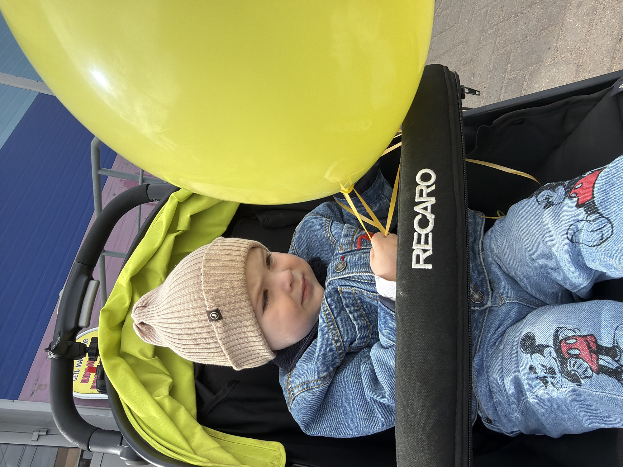 The image shows a baby in a stroller. The baby is wearing a beige beanie, a denim jacket, and denim pants with a Mickey Mouse print. The stroller has a black frame and is lined with green fabric. A large yellow balloon is positioned near the baby, who is holding the string. The stroller has a "RECARO" logo on the front bar. The background shows a building with blue and gray siding. The exact location in Votkinsk, Russia, cannot be determined from the image alone.