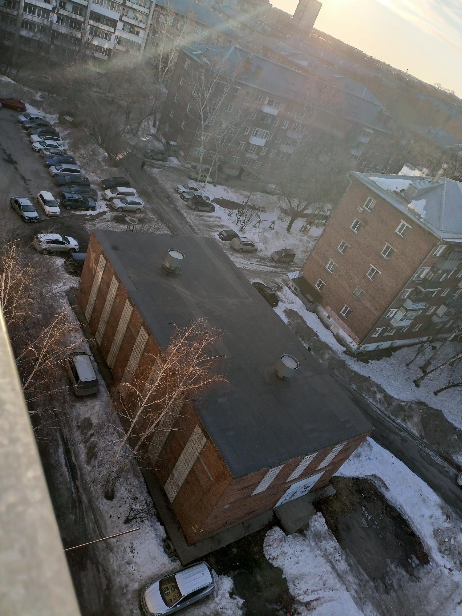 The image shows an aerial view of a neighborhood in Novosibirsk, Russia, possibly taken from an upper floor of a residential building. The scene includes several apartment buildings, with a prominent brick building in the foreground, distinguished by its flat roof and rectangular shape. Cars are parked along the streets and in designated parking areas, some covered with snow. The environment suggests a winter or early spring season, as patches of snow and ice are visible on the ground and on the roofs of buildings. The overall tone is muted with a hazy or overcast sky.