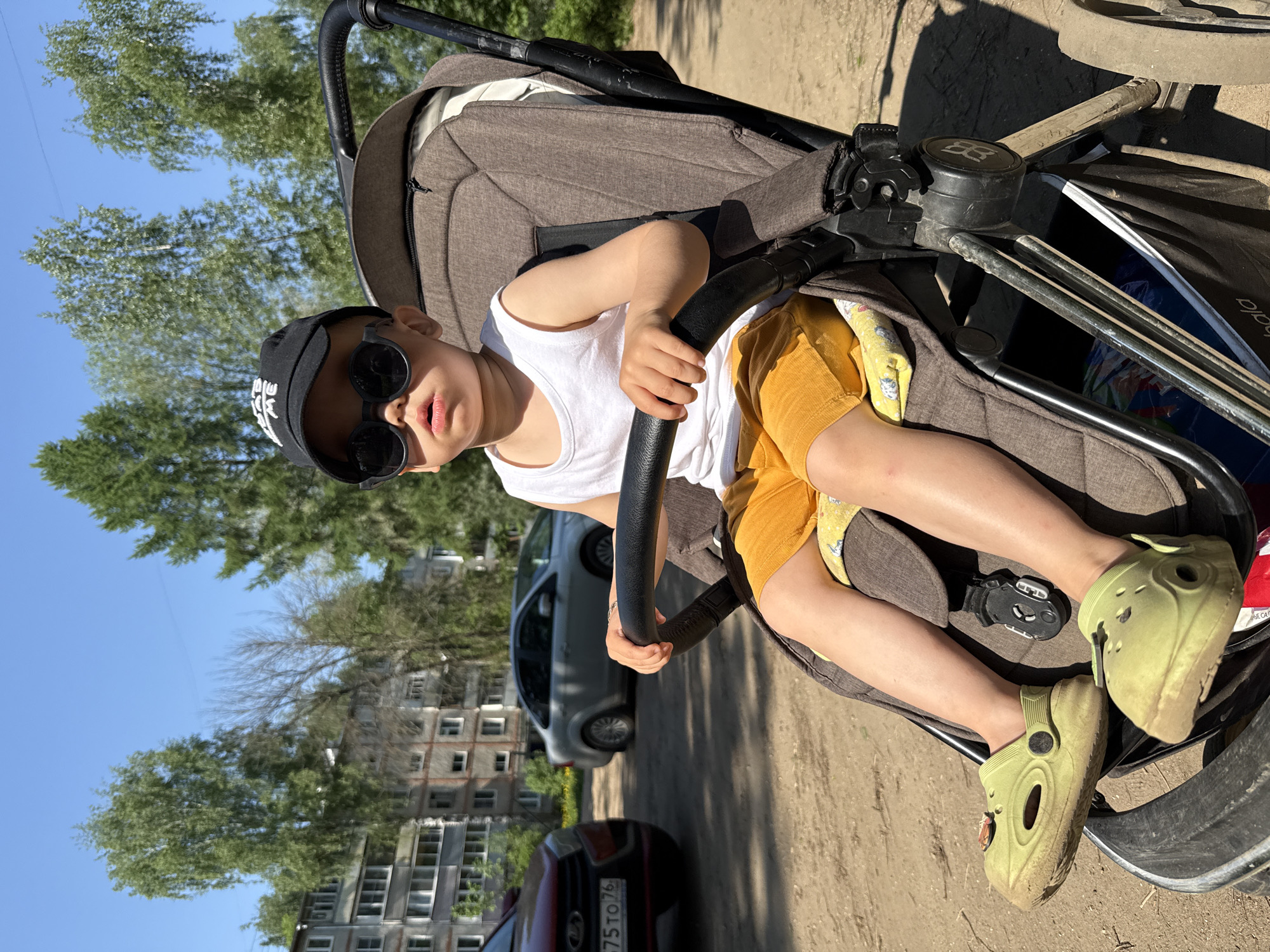A child wearing a white tank top, yellow shorts, a black baseball cap, and sunglasses is seated in a brown stroller. The stroller is positioned on a dirt surface with buildings and trees visible in the background. Cars are parked nearby. The scene is outdoors during daylight hours. The location is Rusanovo, Russia.