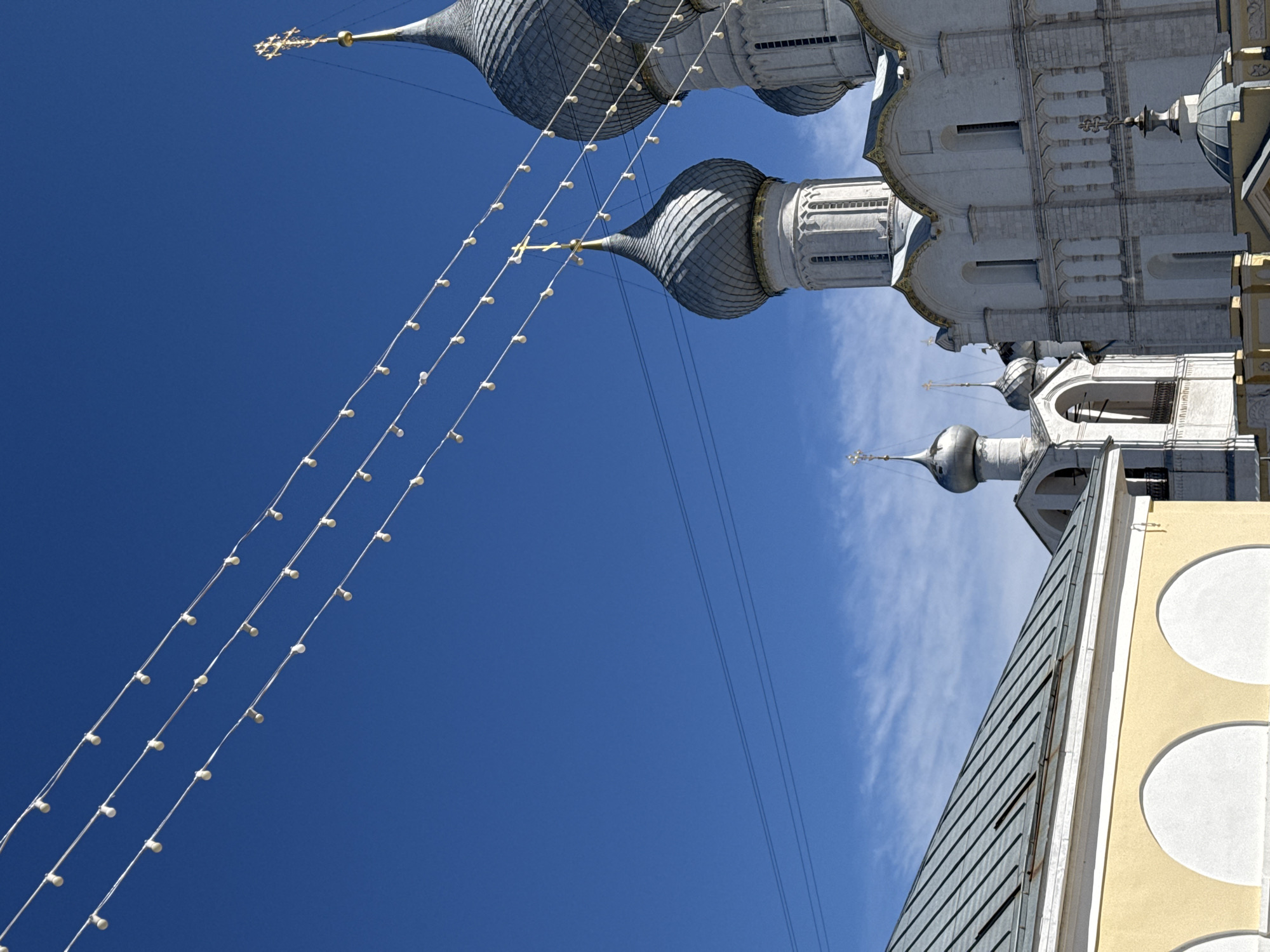 The image depicts a striking architectural scene, dominated by several grey, ribbed onion domes characteristic of Orthodox Christian churches, set against a brilliant clear blue sky. The domes, some topped with golden crosses, rise from white stone structures detailed with arched windows and decorative elements, hinting at a church or monastery complex.
Three prominent strands of string lights, adorned with numerous small white bulbs, stretch diagonally across the upper and central parts of the sky, adding a decorative or festive touch. Additionally, thin power lines are visible, traversing the clear sky. In the lower right corner, a section of a yellow building with distinctive white circular design elements is partially visible, contrasting with the main white architectural structures.
The bright lighting and deep blue sky indicate a clear, sunny daytime setting, with only a few faint, wispy clouds appearing in the lower right background. There are no people or explicit activities shown. The architectural style strongly suggests a location within Russia, consistent with the provided context of Ново-Никольское. No visible text is present in the image.