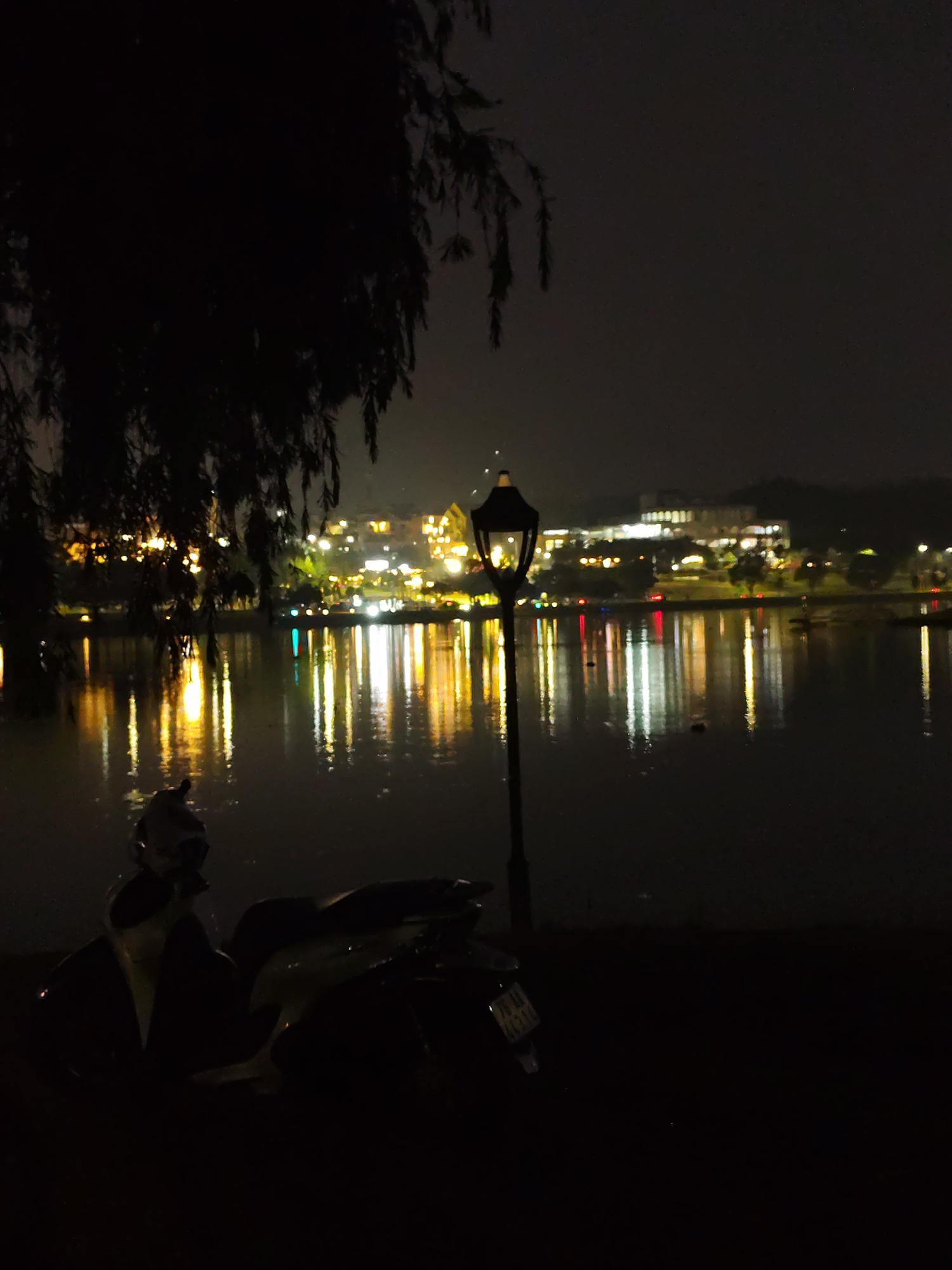 The image depicts a nighttime scene at Xuan Huong Lake in Da Lat, Vietnam. A lake is visible in the center with the reflection of lights on the surface. A vintage-style lamppost stands to the right of the frame. A parked scooter is in the foreground, to the left of the lamppost. Dark silhouettes of tree branches are visible to the upper left of the frame. Buildings with lights on are visible in the background across the lake.