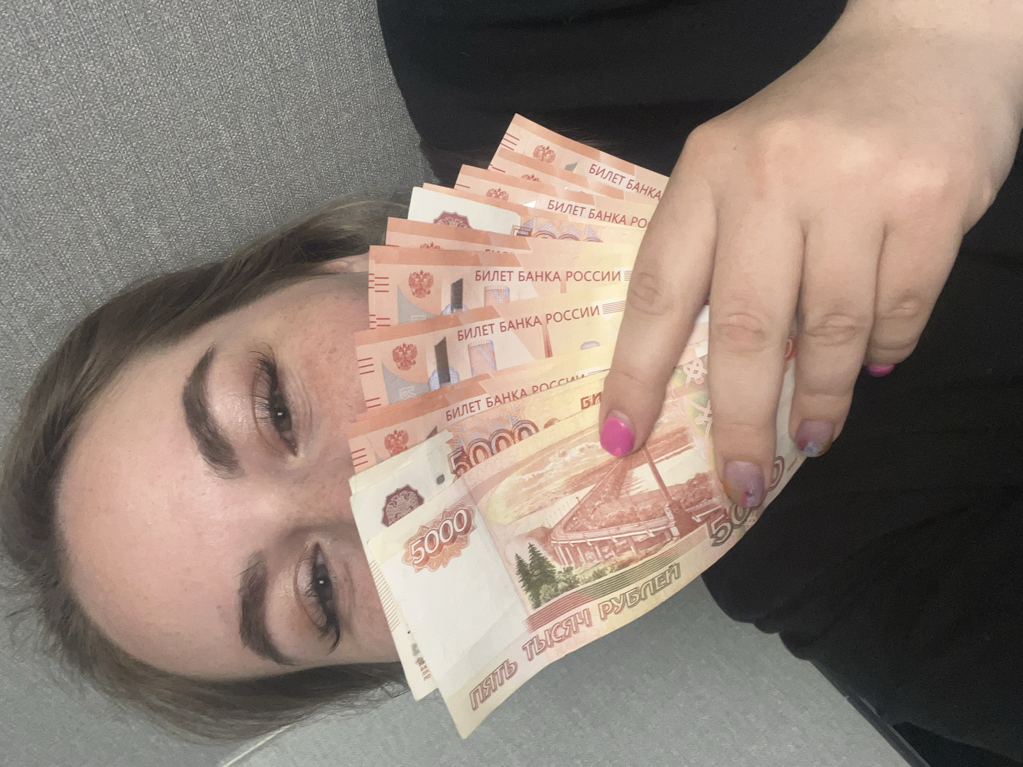 An image taken in Chaikovsky, Russia, features a person lying down and holding a fan of Russian 5000 Ruble banknotes. The photograph is rotated, showing the person's head on the left and their body extending to the right. Visible are the person's upper face, from the forehead to the chin, and their right hand. The individual has groomed eyebrows, long eyelashes, and open eyes that appear to look towards the camera. Freckles are discernible on their fair skin.
The right hand is prominently displayed, holding a stack of several 5000 Ruble banknotes which partially obscure the person's lower face. The thumb features bright pink nail polish, while the other visible fingernails have a multi-colored, possibly iridescent or glittery, finish. The banknotes are predominantly pink-orange and clearly display "5000" as their denomination, along with the text "БИЛЕТ БАНКА РОССИИ" (Bank of Russia Note) and "ПЯТЬ ТЫСЯЧ РУБЛЕЙ" (Five Thousand Rubles). The person is resting on a textured grey surface, likely a sofa or bed, with a darker fabric visible to the right, possibly an item of clothing or another part of the furniture. The scene conveys a casual, indoor setting where a substantial sum of money is being displayed.