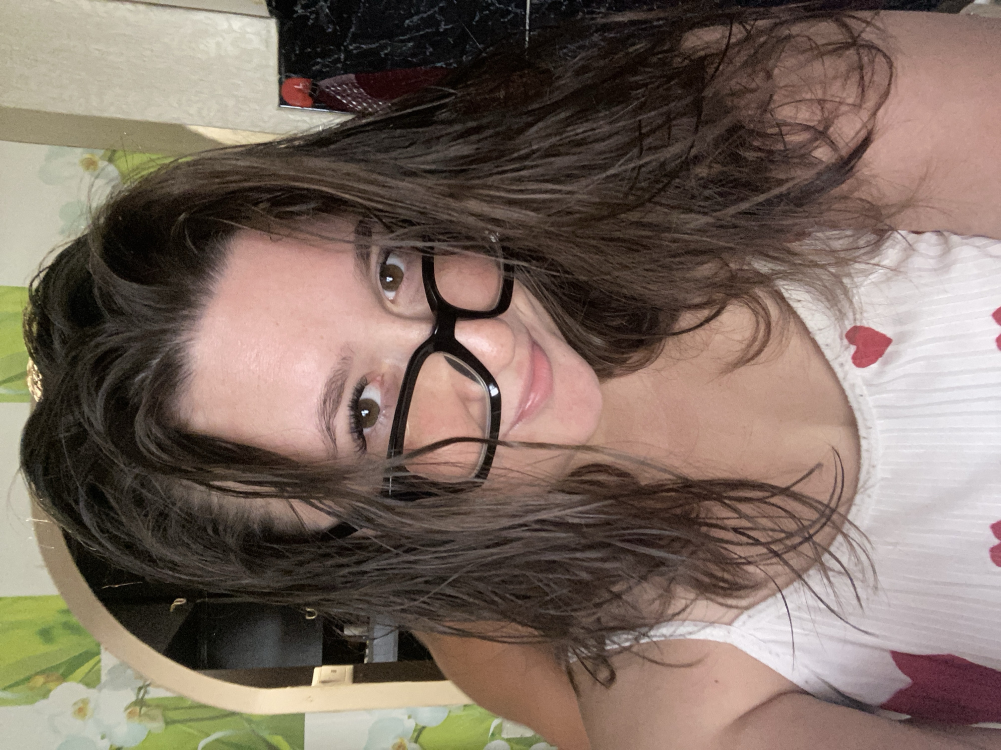 The image shows a close-up selfie of a woman with dark, wet hair and dark-rimmed glasses. She is wearing a white tank top with red heart patterns. Her eyes are brown, and she is smiling slightly. The background is out of focus but appears to be an indoor setting with floral wallpaper and a dark, possibly mirrored, surface. There is no visible indication of the city of Chaikovsky, Russia.