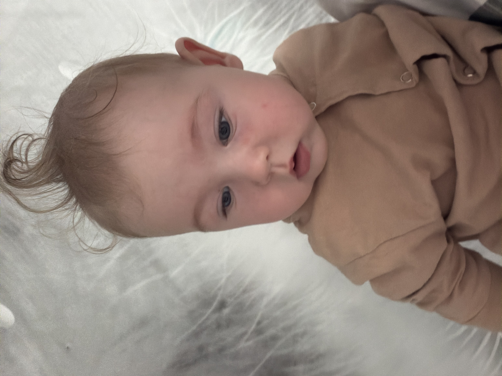 An extreme close-up shot focuses on the face and upper body of a baby lying down, viewed from a high angle. The baby has light skin, blue eyes, and sparse light brown hair styled into a wispy tuft on top of its head. It is wearing a tan-colored onesie with silver snaps. The background consists of a textured white surface, possibly bedding, with soft lighting. The image does not provide any information about the city of Chaikovsky, Russia, or any specific location.