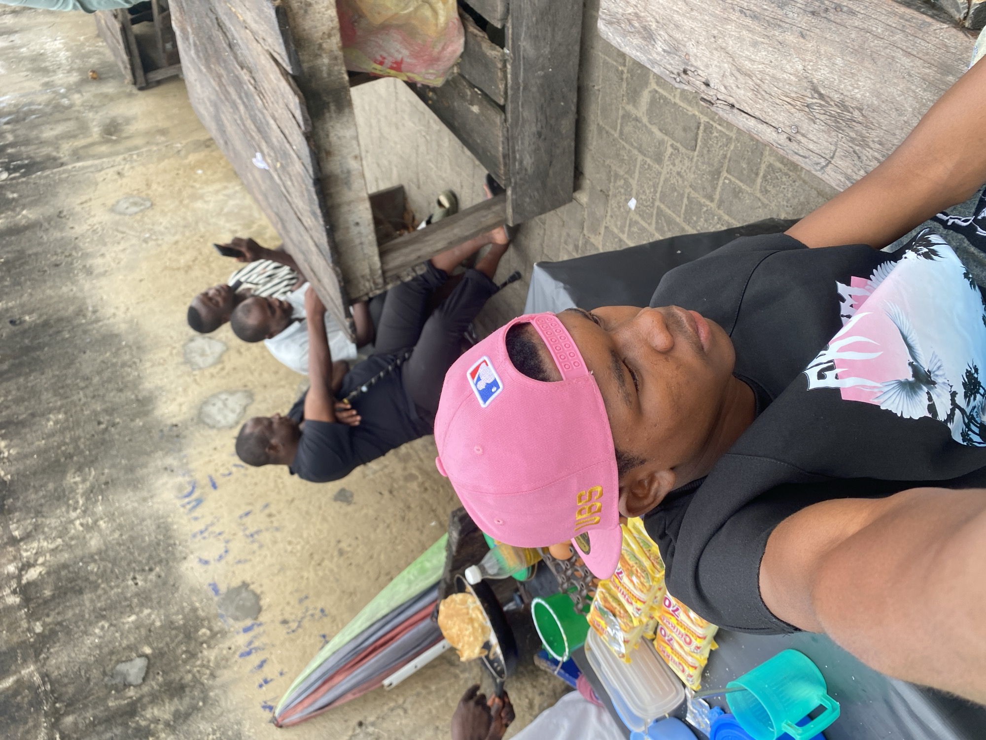 In an outdoor urban setting, likely a street market or roadside food stall, several individuals are gathered. In the foreground, a young person wearing a pink baseball cap and a black t-shirt with a graphic print is captured in a selfie-style shot, looking downwards. To their left, on a black surface, are several packets of instant noodles, a blue plastic cup with a spoon, and a white container. Further back, two men are seated at a wooden table. One man, wearing a black t-shirt, is partially obscured, while the other, in a white and striped shirt, is holding a mobile phone and looking at it. Behind them, another man in a black t-shirt is seated, his arms crossed. To the left of the frame, an umbrella with green, red, and gray panels is propped up, and a hand is visible cooking food in a pan. The background features a textured, weathered concrete wall with some blue markings. The ground is paved with interlocking bricks.
