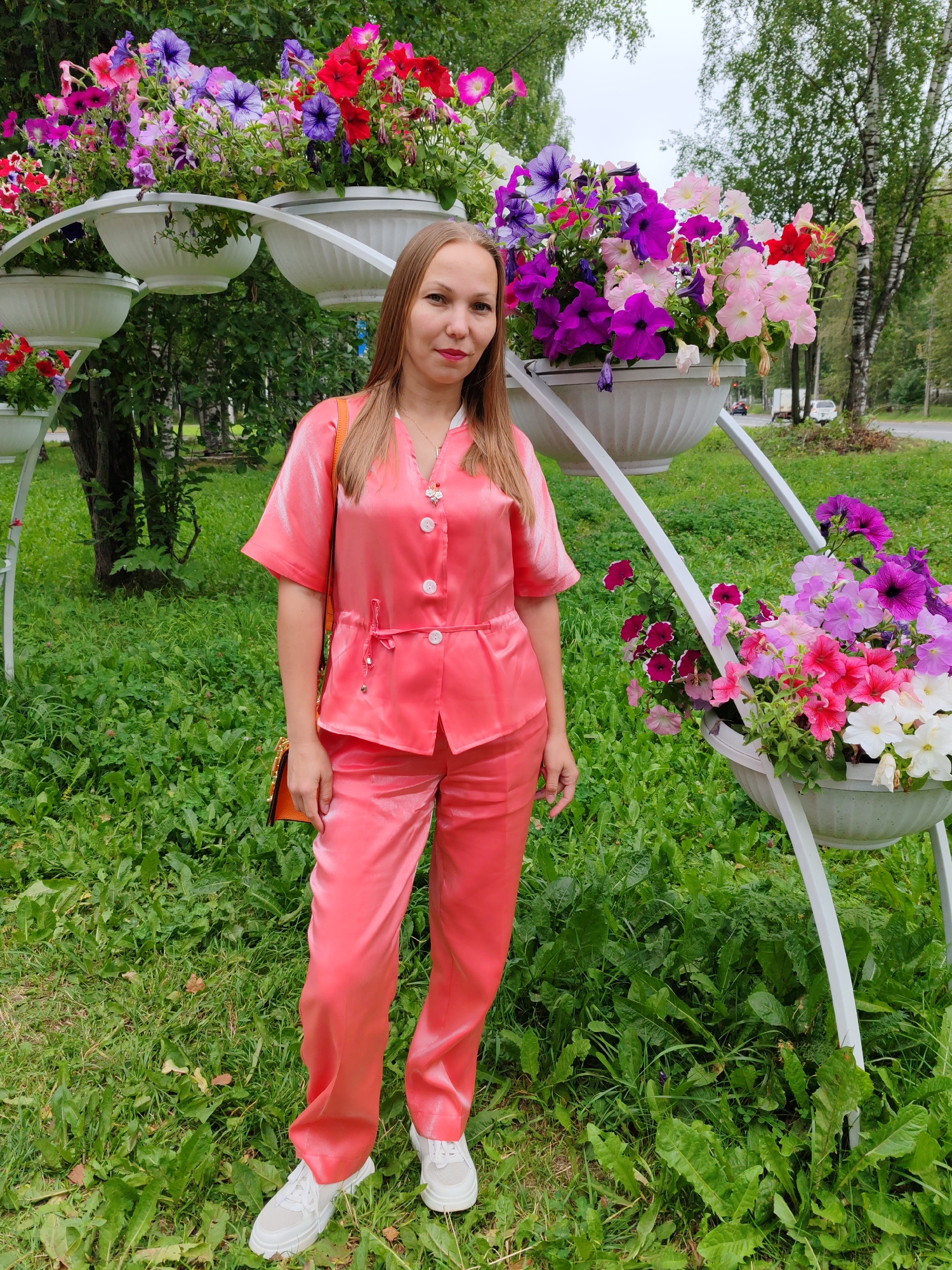 A young woman with long blonde hair stands in a park in Syktyvkar, Russia. She is wearing a coral-colored silk pajama set with a V-neck top and matching pants. A small orange purse hangs over her left shoulder. She is wearing white sneakers. The woman is standing in front of a white tiered planter filled with vibrant pink, purple, and red petunias. The background shows green grass, trees, and a street with a car and a truck visible. The sky is overcast.