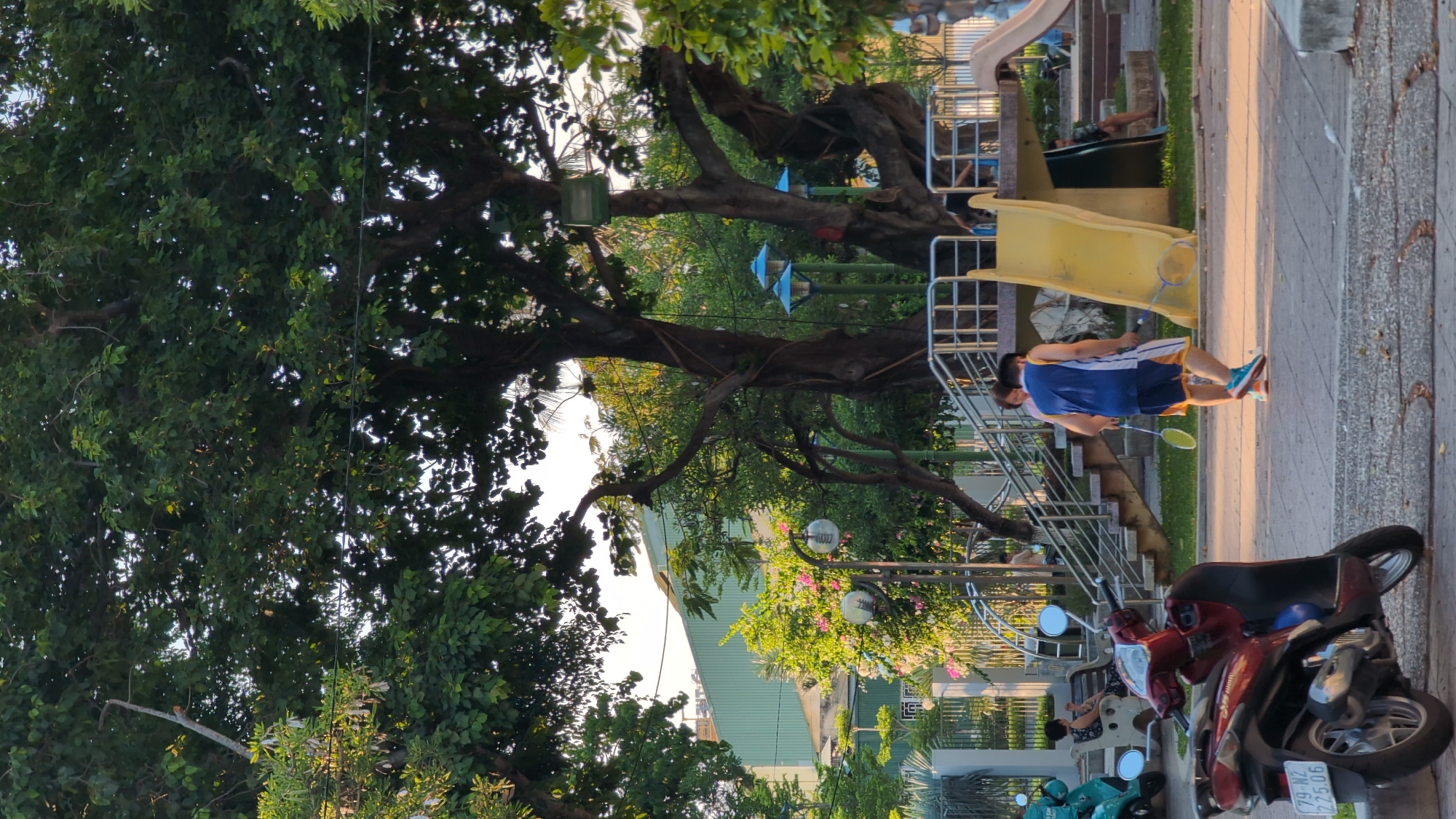 The image depicts an outdoor scene in a public park, likely in the city of Nha Trang Ward, Vietnam. The foreground is dominated by a red motorcycle parked on a paved surface. Beyond the motorcycle, a playground area is visible, featuring a yellow slide and stairs. Two individuals, a man and a woman, are engaged in a game of badminton near the playground. The man is wearing a blue and white sleeveless shirt and blue shorts, while the woman is in a pink top. In the background, a large tree with dense foliage provides shade. Further back, a light green building and a fence are discernible. Several other people are present in the park, some sitting on benches and others in the background, contributing to the impression of a typical day in a community space. The overall atmosphere suggests a relaxed, daytime activity.