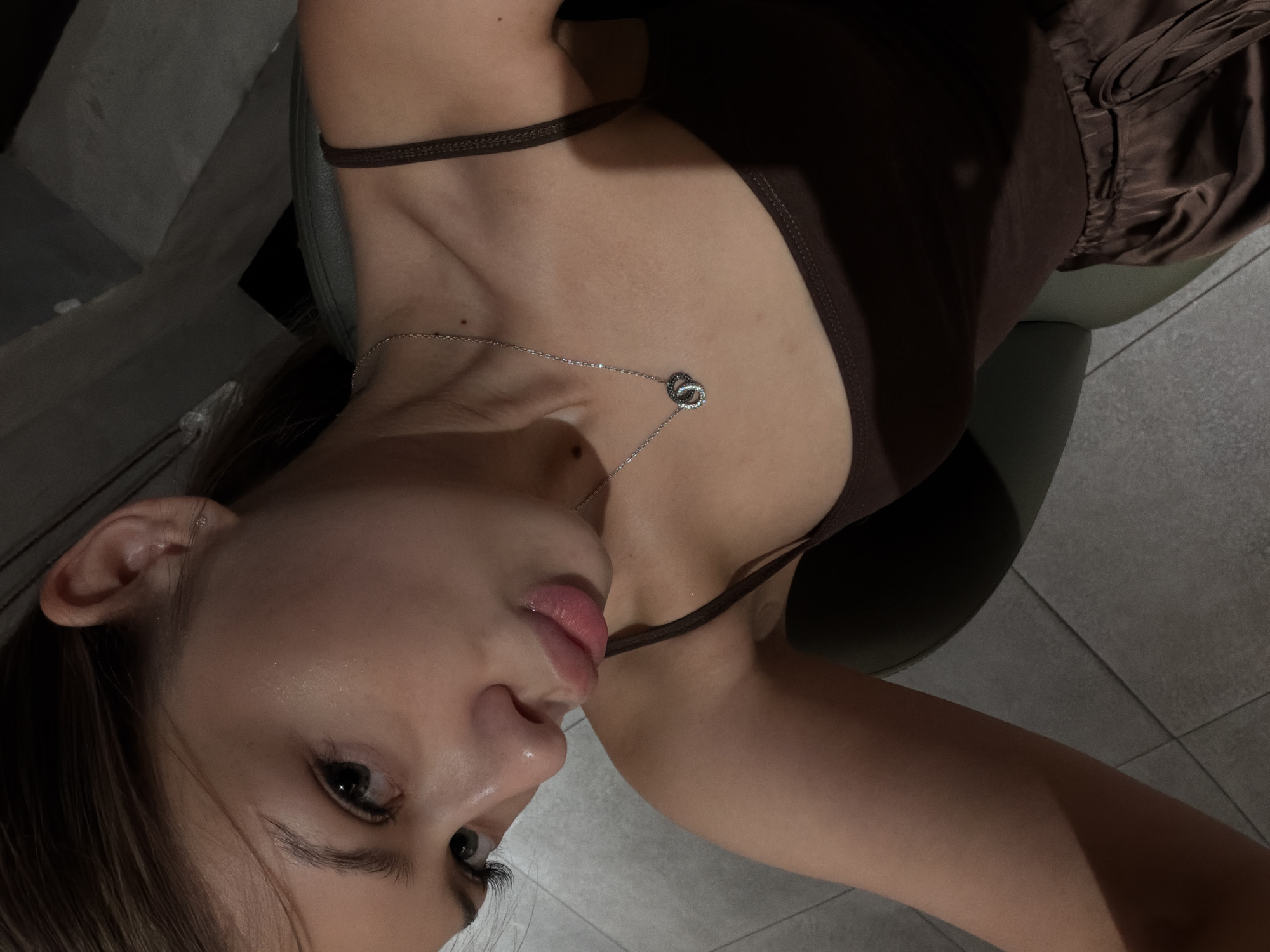 A young woman is captured in a close-up selfie, with her head tilted downwards and to her right. She has dark hair pulled back, and her face is illuminated, highlighting her features, including her eyes, nose, and full lips which are slightly pursed. She is wearing a dark brown spaghetti strap top and a silver necklace with two interlocking rings. The background appears to be indoors, with a tiled floor and a dark, indistinct area behind her. The lighting suggests a low-light environment, possibly a home or a dimly lit room. There is no indication of Đà Nẵng, Vietnam in the image.