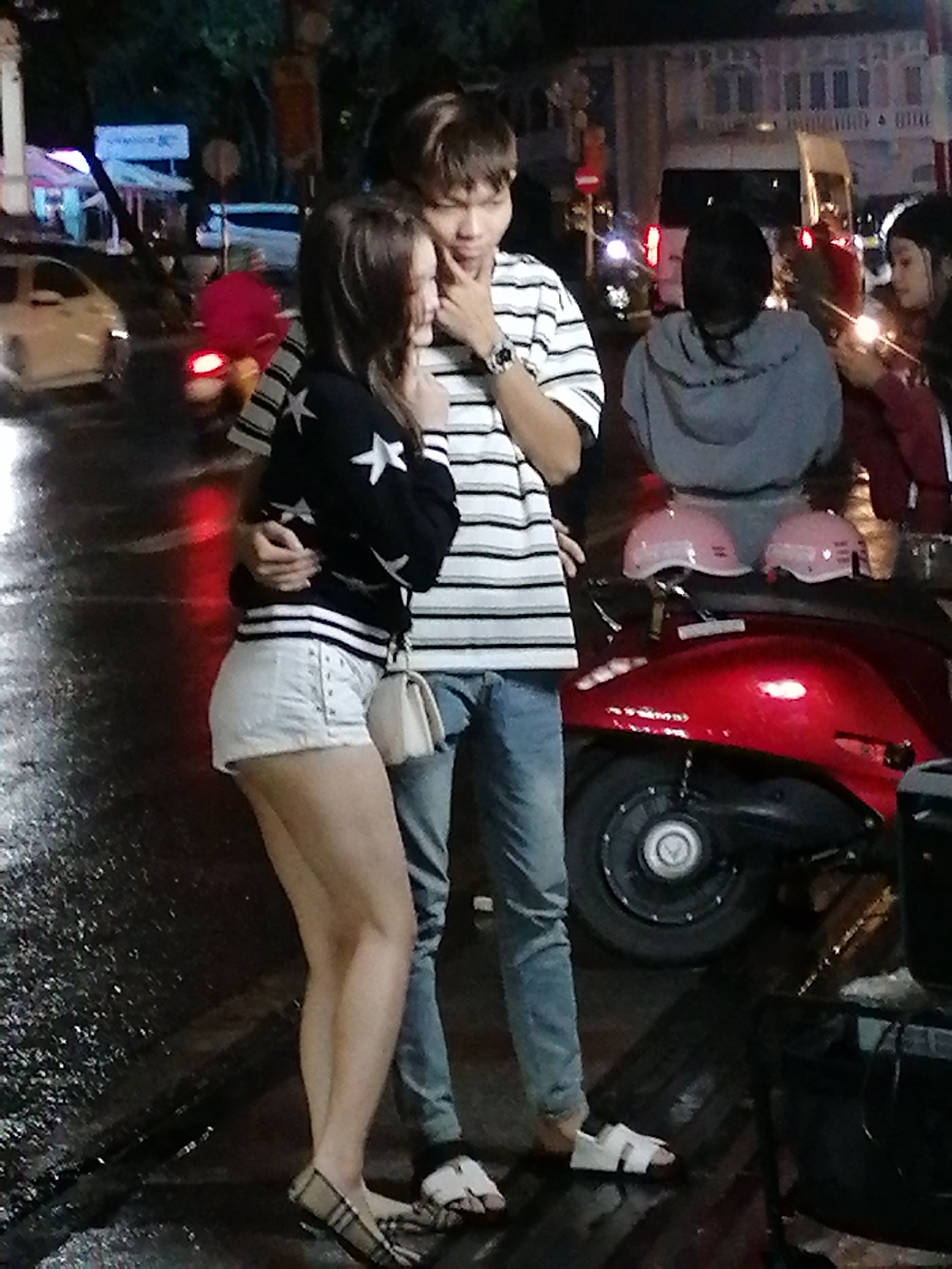 A young Vietnamese couple is standing on a wet city street at night, possibly in Da Lat, Vietnam. The woman, wearing a black sweater with white stars and white shorts, is embracing the man. The man, in a striped t-shirt and jeans, has his arm around her. A red scooter with two pink helmets is parked behind them. The street appears to be illuminated by streetlights and the lights from nearby buildings and vehicles, creating reflections on the wet pavement. Other people and vehicles are visible in the background, suggesting a bustling urban environment.