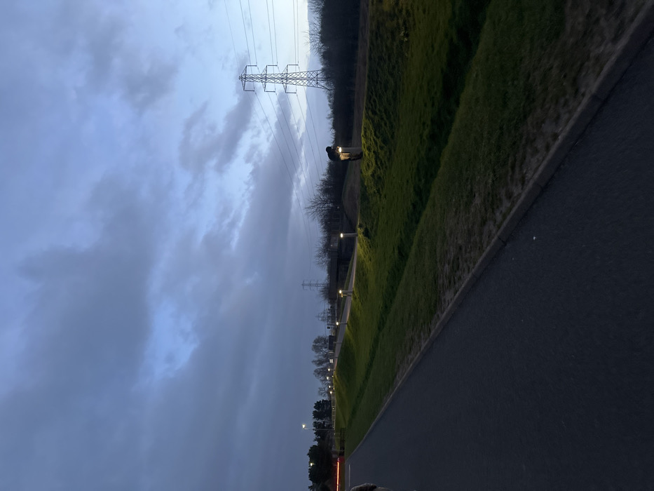 The image depicts an outdoor scene in Falkirk, United Kingdom, at dusk or early evening, characterized by low natural light and active artificial illumination. The sky is largely overcast with dark grey clouds, transitioning to a lighter blue on the right horizon.
In the foreground, a dark asphalt pathway extends from the lower left towards the center-right. The edge of a human figure in light-colored clothing is partially visible on the far left of this path. Adjacent to the path on the right, a grassy embankment slopes upward, bordered by a drainage ditch. Along the crest of this embankment, a series of evenly spaced, illuminated bollard lights cast circular pools of light onto the grass.
Further into the midground, beyond the first embankment, another paved area or pathway is visible, also lined with similar bollard lights. A lone individual, dressed in dark clothing, is positioned on the first grassy embankment, leaning forward and appearing to interact with or examine one of the illuminated bollard lights on the right side of the frame. Their posture involves bending at the waist over the light fixture.
The background features a line of bare deciduous trees and shrubs. Two large metal lattice utility poles (pylons) with overhead power lines are visible; a prominent one on the far right and a smaller one to its left. A dark bridge structure crosses the scene horizontally in the mid-left background. Distant, indistinct artificial lights are visible on the far left horizon.