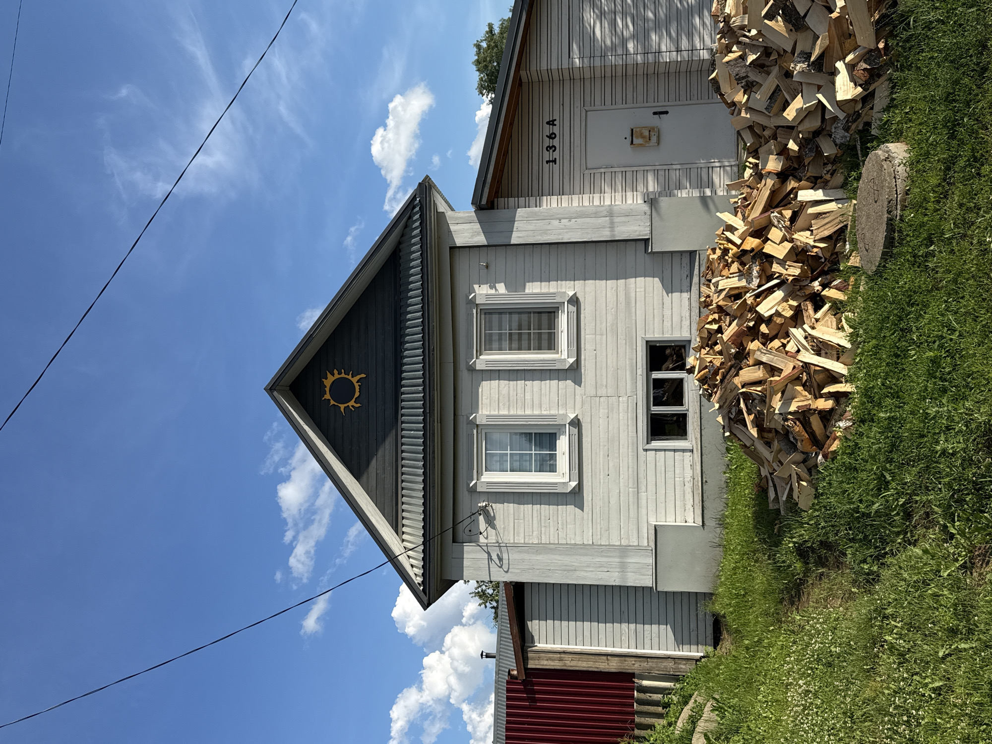 This media file depicts a wooden house with a pitched roof in Votkinsk, Russia. The house is painted light gray, and its gabled end features a dark gray section with a decorative yellow sun emblem. Two windows are visible on the front of the house, one above the other, both with white frames. Above the upper window, the house number "136A" is visible in black lettering. To the right of the house, a large pile of chopped firewood is stacked high against the wall.
The scene is set outdoors under a bright blue sky with scattered white clouds. The sun is shining, suggesting it is daytime. The presence of firewood indicates a preparation for cold weather, or that the house is used as a primary residence where heating with wood is common. Lush green grass and some wildflowers grow around the base of the house, suggesting a rural or suburban setting in a temperate climate. There is a metal fence visible at the bottom of the frame, painted red, indicating a boundary. Electrical wires are also visible traversing the sky. The overall impression is one of a simple, functional home in a natural environment. No people are visible in the image.